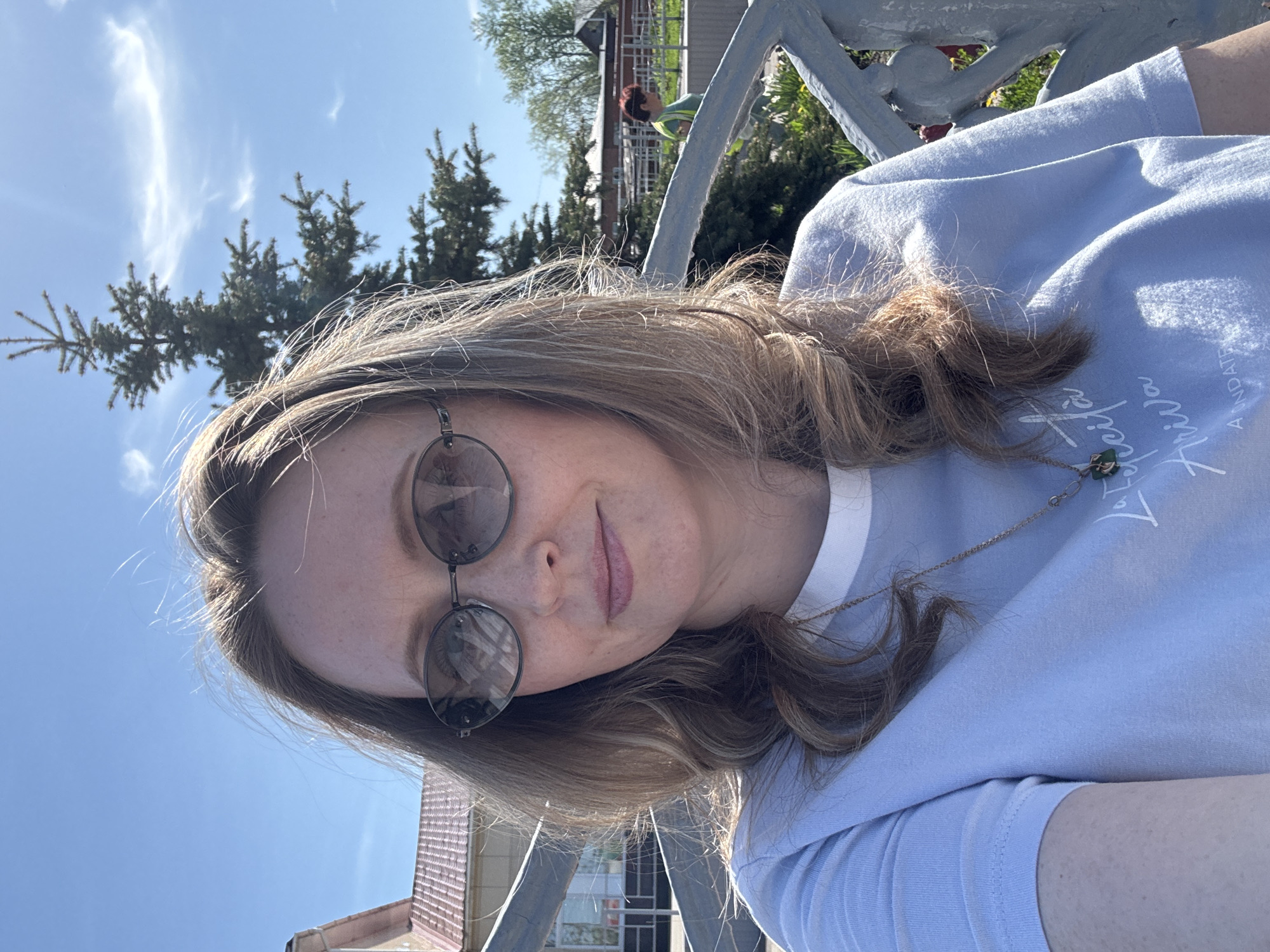 A woman with shoulder-length brown hair and sunglasses is sitting on a gray metal bench outdoors. She is wearing a light blue t-shirt with white lettering. Behind her, there is a blue sky with some clouds, as well as some trees. In the background, there is a building with a brown roof. Based on the surrounding environment, this could be in Balezino, Russia. A person is walking in the background.