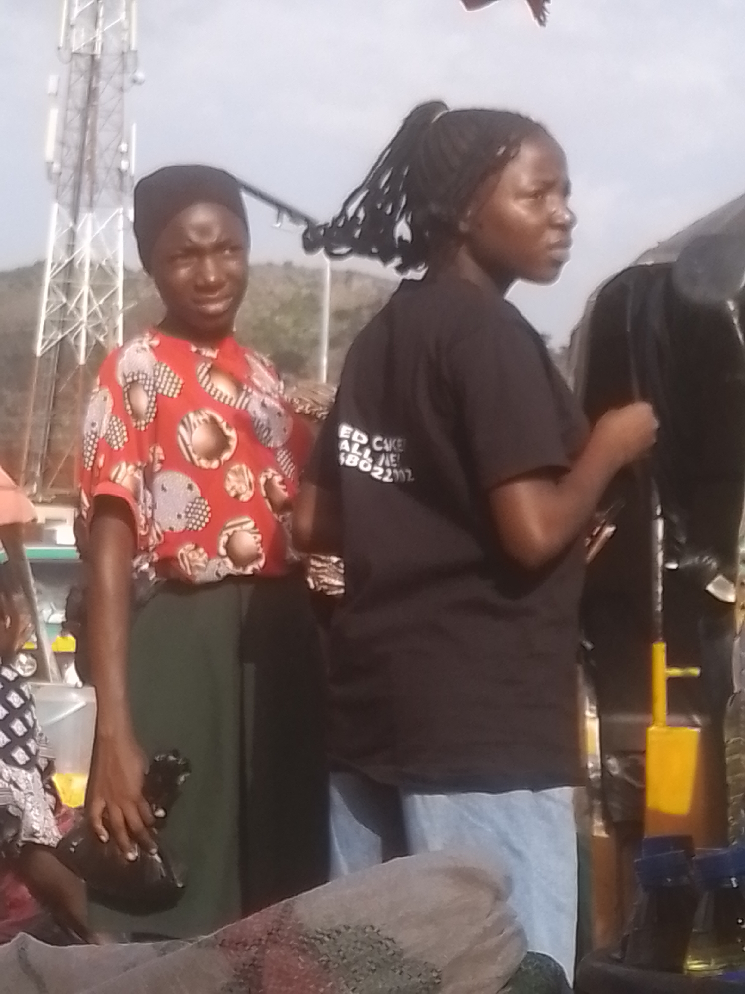 The image depicts two young women standing outdoors. The woman on the left wears a black headscarf and a red shirt with a white, brown, and black pattern. She holds a dark, plastic-wrapped object in her right hand. The woman on the right has her hair in braids pulled back into a ponytail and wears a black t-shirt with white text on the back. She is looking to her right. Behind them, a tall cell tower is visible against a bright, overcast sky. The background includes a hint of hilly terrain. The overall lighting suggests it is daytime, possibly late afternoon due to the soft light. In the foreground, various objects are partially visible, including what appears to be a mesh bag and some bottles. The scene appears to be a public space, possibly a market or roadside, given the presence of the tower and the various items arranged around. The facial expressions of the women are neutral, and there is no clear indication of a specific event or activity beyond them standing together. Text is visible on the t-shirt of the woman on the right, which appears to read "ED CAKE ME 2202".