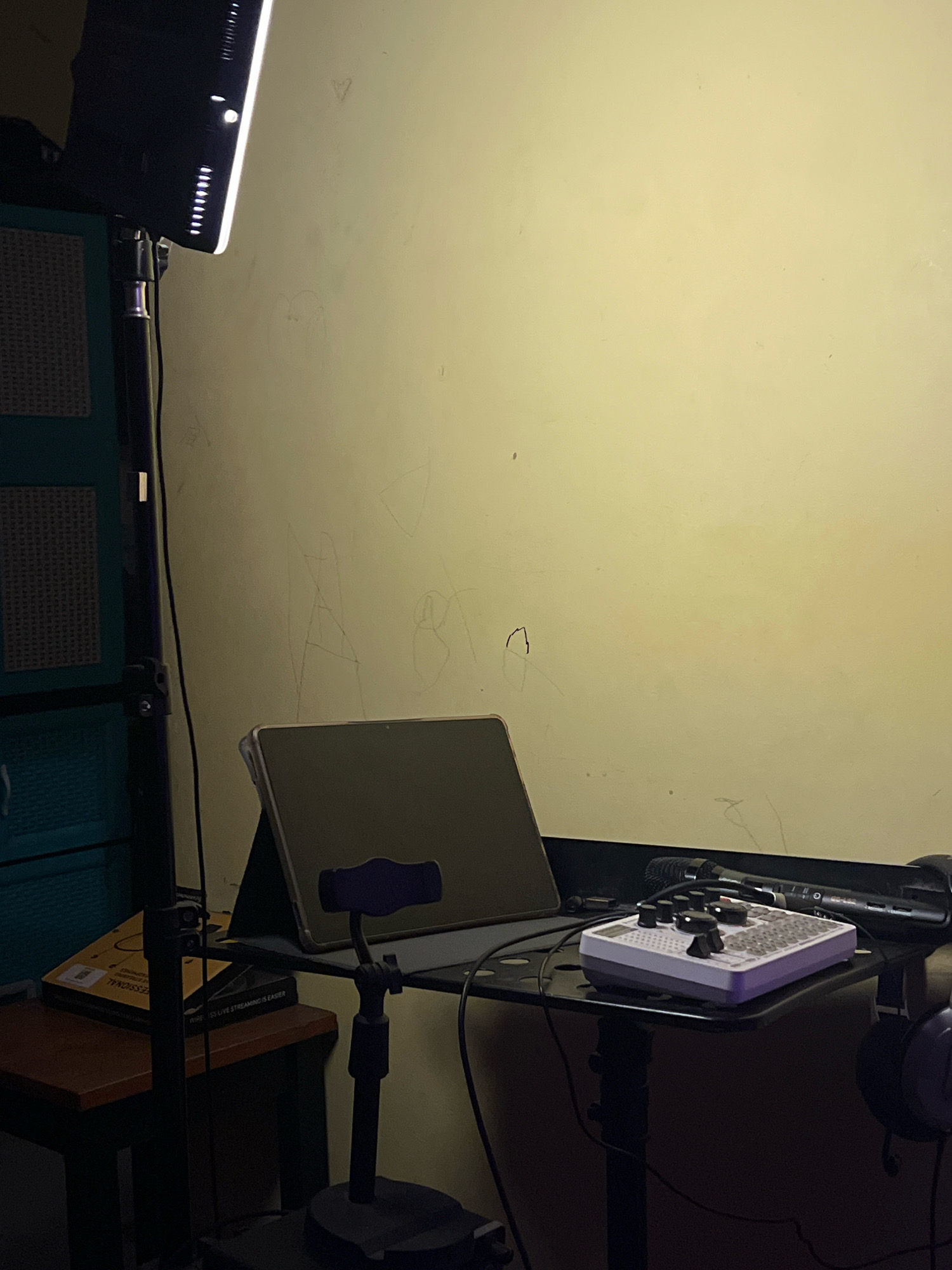 The image depicts a desk setup for content creation, likely in a bedroom or small studio in Sleman, Indonesia. The scene is dimly lit, with a large LED light panel on a stand dominating the left side of the frame. The light casts a warm glow on a pale yellow wall, which has faint, scribbled drawings on it.
On the desk, a tablet is propped up in a black stand, its screen dark. To the right of the tablet sits a small, white audio interface with several knobs and buttons. A black microphone is positioned behind the audio interface, and a pair of purple headphones rests on the far right edge of the desk. Cables are visible, connecting the various devices.
To the left of the desk, a small wooden table holds a book with a yellow cover, partially obscured by a black microphone stand. The background features a portion of a teal cabinet with a textured surface. The overall impression is of a functional, albeit somewhat cluttered, creative workspace.