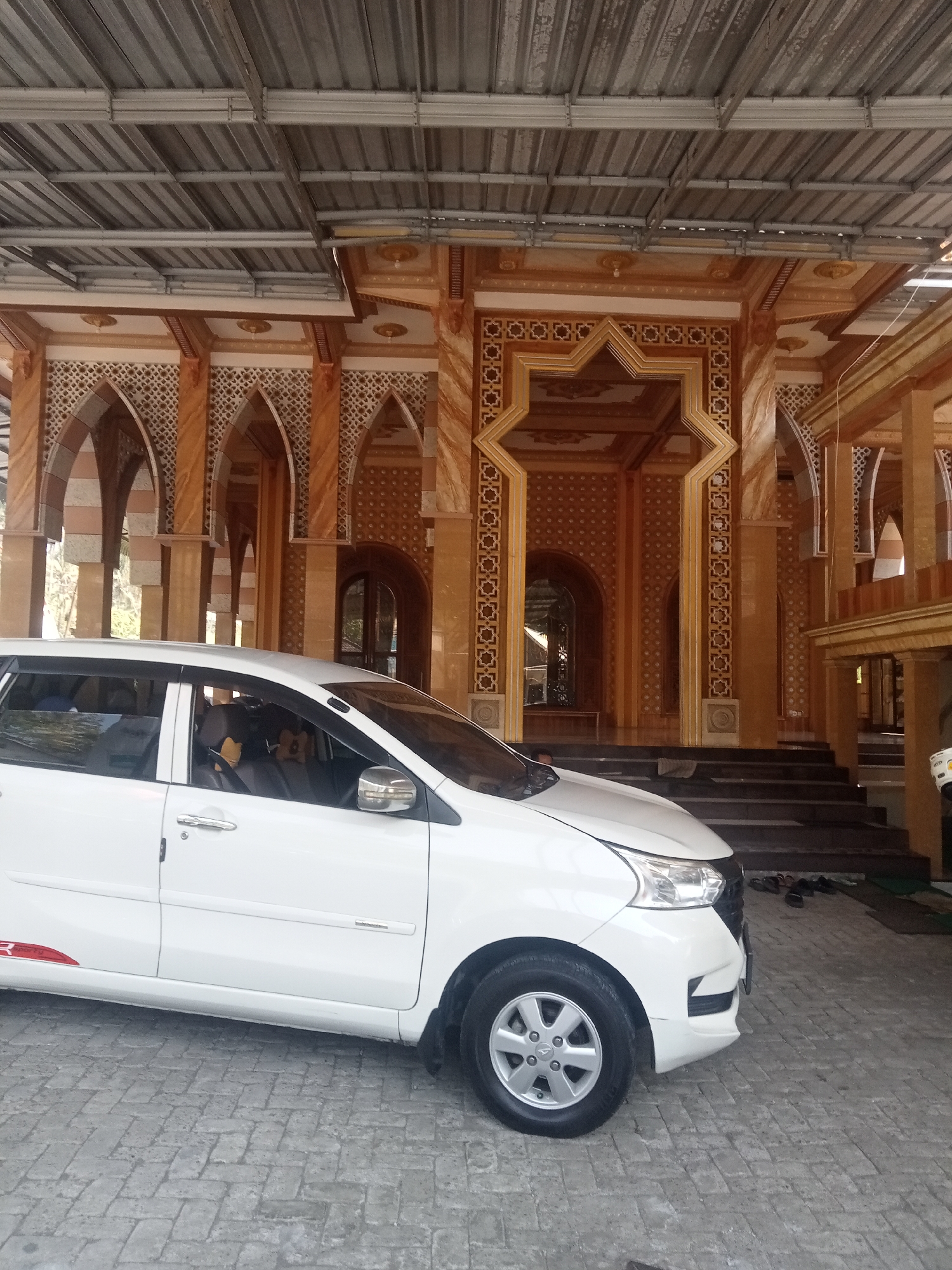 A white car is parked on a stone-paved surface in front of a building with ornate architecture, likely a mosque, in Tlogosari, Indonesia. The building features intricate geometric patterns and arched doorways with marble-like columns. The car's driver's side is visible, showing its door, side mirror, and front wheel. Several pairs of sandals are placed on a dark mat near the building's steps. A metal roof structure with exposed beams is visible above the building's facade. The scene is outdoors with natural light.
