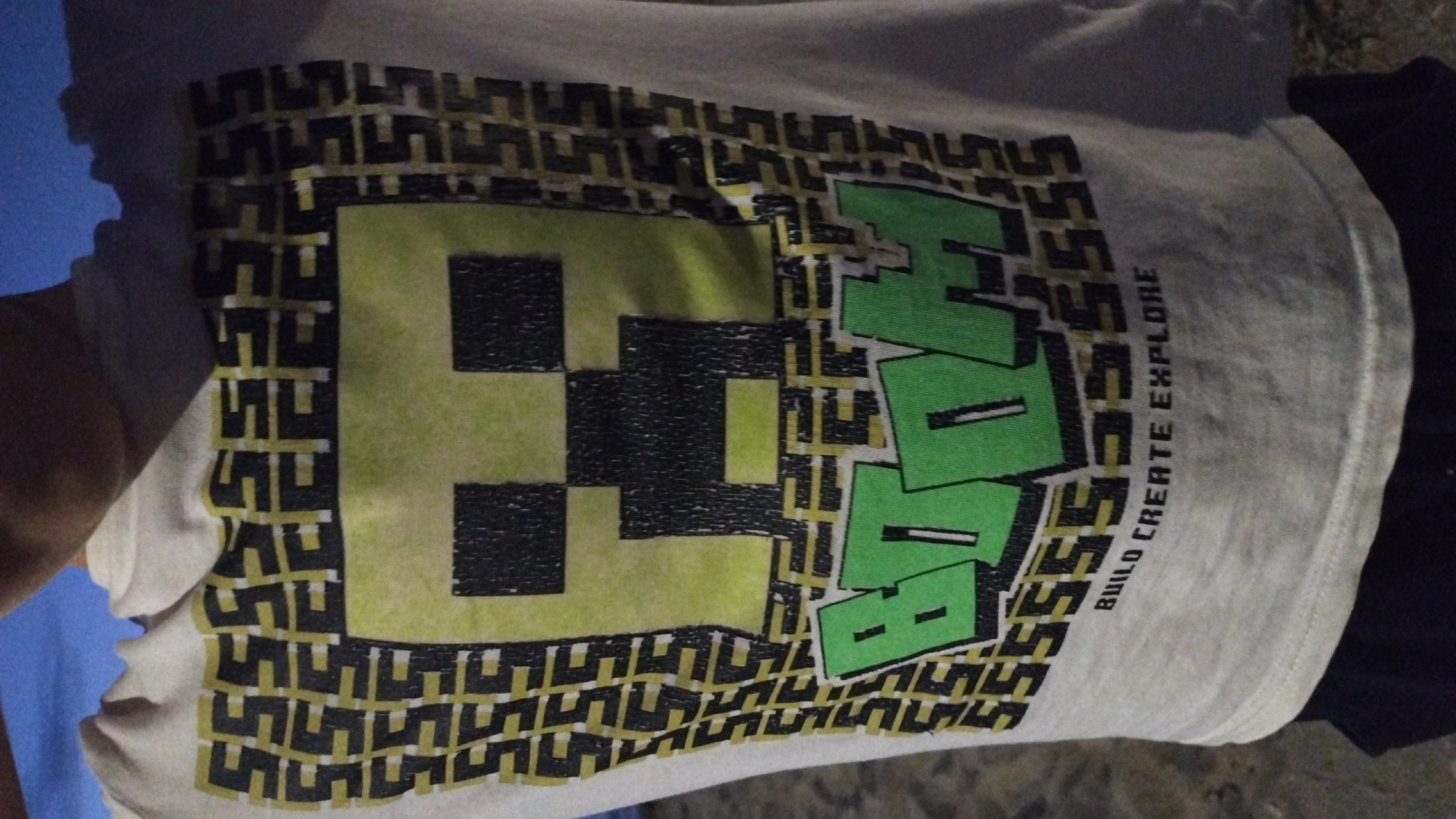 A close-up, slightly tilted shot shows a cream-colored t-shirt with a graphic design. The graphic features a pixelated green and black creeper face on the left, a large green word that appears to be "HIVE" on the right, and a repeating pattern of black and yellow shapes forming a border around the design. Below the "HIVE" text, in smaller vertical lettering, are the words "BUILD CREATE EXPLORE." The t-shirt is being worn, and the wearer's arm and shoulder are partially visible on the left side of the frame, along with a hint of a blue garment underneath. The t-shirt is laid out on a dark surface, possibly a floor or ground, with a dark blue or black garment visible beneath the right side of the t-shirt. The lighting is dim, suggesting it is nighttime or indoors. The overall impression is of a casual, possibly gaming-themed t-shirt. There are no explicit clues about the time of day, weather, or specific location within Perea, Greece, beyond the provided context. The image does not convey any discernible emotions.