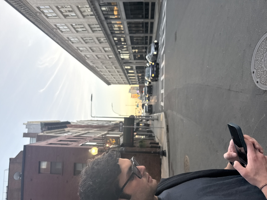 The image depicts an urban street scene in the downtown area of Boston, Massachusetts, United States, during what appears to be late afternoon or early evening.
In the foreground, slightly left of center, a male individual with dark, curly hair and dark sunglasses is seen in profile, looking down at a dark-colored smartphone held in both hands. He wears a dark t-shirt and a dark open jacket or vest. A light-colored bracelet is visible on his left wrist. Part of a circular metal manhole cover with a geometric pattern is visible on the asphalt pavement in the lower right.
The street, paved with asphalt, extends into the background, showing white crosswalk markings in the mid-ground. Multiple cars are present on the street, traveling away from the viewer; some have illuminated headlights. The street is flanked by multi-story buildings. On the left, a red-brick building with visible windows features a prominent street lamp with a glowing yellow light and a "ONE WAY" sign below it. Some decorative, warm-toned lighting is visible near an entrance on this building. On the right, a lighter-colored, possibly limestone or light brick, multi-story building with numerous large windows on its lower floors and smaller windows on upper floors lines the street, reflecting the sky and interior lights.
The sky in the background is light blue with a warm, yellowish glow near the horizon, and faint white contrails are visible. Distant urban architecture and an illuminated sign are discernible further down the street.