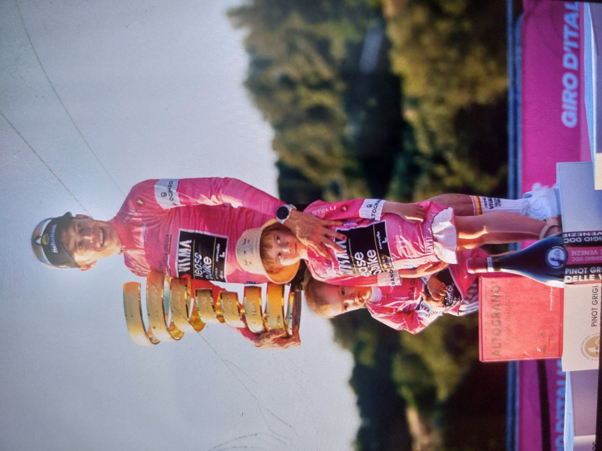 The image shows a cyclist, presumably a race winner, posing with two children, all dressed in pink cycling outfits with "VISMA | Lease a Bike" logos. The cyclist holds a multi-tiered gold trophy. The background features green foliage and a pink "GIRO D'ITALIA" banner, suggesting this is a Giro d'Italia victory celebration. Additional items such as wine bottles and boxes, including a box labeled "ALTOGRANO", are present on a table or platform next to the cyclist. The location is not Tsarevo, Bulgaria, but rather somewhere associated with the Giro d'Italia cycling race.