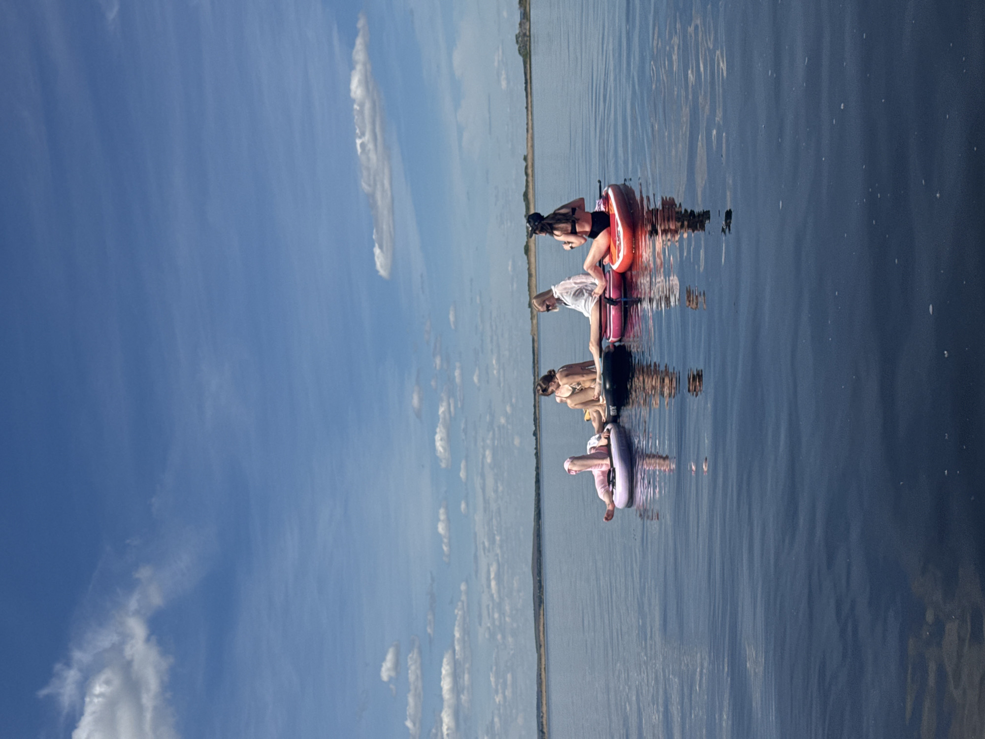 The image captures a serene outdoor scene, likely on a large body of water, with several individuals enjoying a leisurely day.
**Subjects:** The primary subjects are four people, predominantly women, and one small child. They are seated on or floating beside various inflatable tubes or rafts. The visible rafts include an orange one, a purple one, and a black one, along with another light pink or purple one for the child. The people are dressed in swimwear, with one woman wearing a black swimsuit and a cap, another in a white shirt and shorts, and a third in a light-colored bikini. The child is dressed in pink full-body attire. The vast expanse of calm water occupies the middle and right portion of the image, while a clear blue sky with scattered white clouds fills the left. A faint, low-lying distant shoreline separates the water from the sky.
**Activity or event taking place:** The individuals are engaged in relaxing and floating on the water using inflatable devices. They appear to be enjoying the tranquility of the surroundings, possibly swimming or simply sunbathing.
**Notable details:** The scene depicts a bright, sunny daytime setting, evidenced by the clear blue sky and the strong, distinct reflections of the people, their rafts, and the clouds on the calm water surface. The water is exceptionally still, creating a mirror-like effect that duplicates the sky and the subjects. The weather is clear with only a few wispy clouds. The distant shoreline suggests a lake or a wide river, like the Volga River, consistent with the location context of Усть-Курдюм, Russia. The overall mood conveyed is one of peaceful relaxation and enjoyment of nature. There is no visible text in the image.