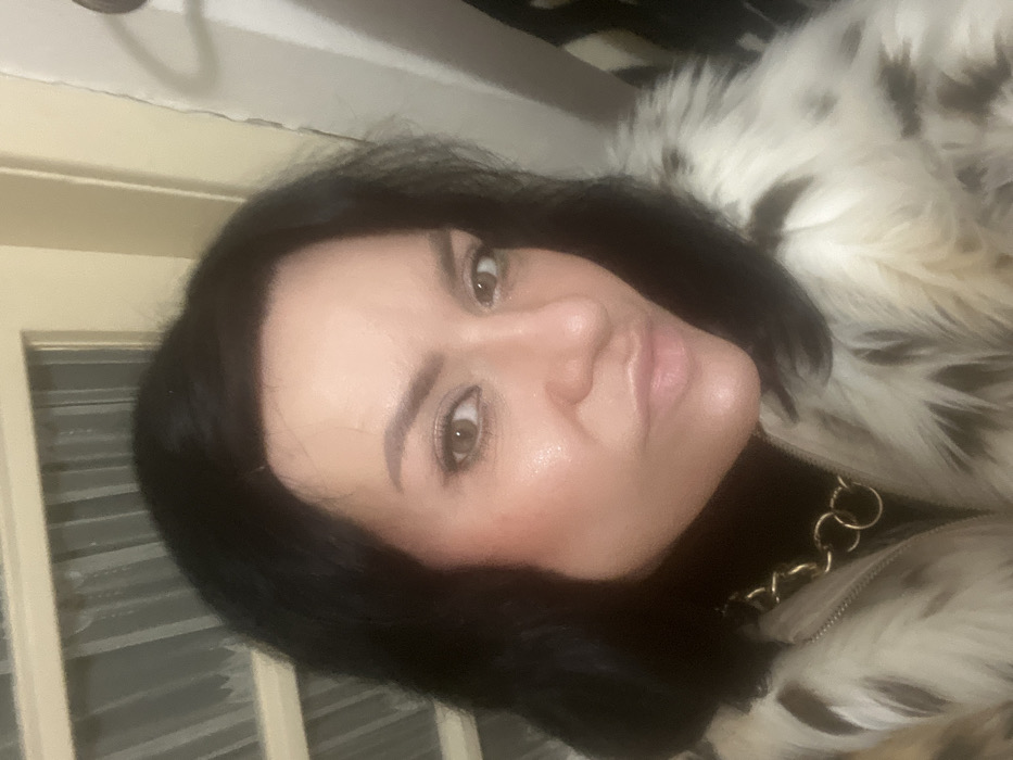 A head-and-shoulder portrait of an adult female, taken indoors in the city of Smederevska Palanka Municipality, Serbia. The subject has dark hair and light brown eyes, looking directly forward. Her facial features include defined eyebrows, prominent cheekbones with blush, and her lips are pursed. She is dressed in a black turtleneck top layered under a coat with a white fur-like texture and irregular black or dark brown patches. A thick, gold-colored chain necklace composed of large circular links is worn over the turtleneck. The background on the left consists of a light-colored, possibly wooden, door or cabinet featuring vertical paneling and a translucent, textured material behind it. The background on the right is a light-colored wall with a vertical seam or crack, and an indistinct dark object is partially visible in the upper right. The scene is illuminated by artificial light.