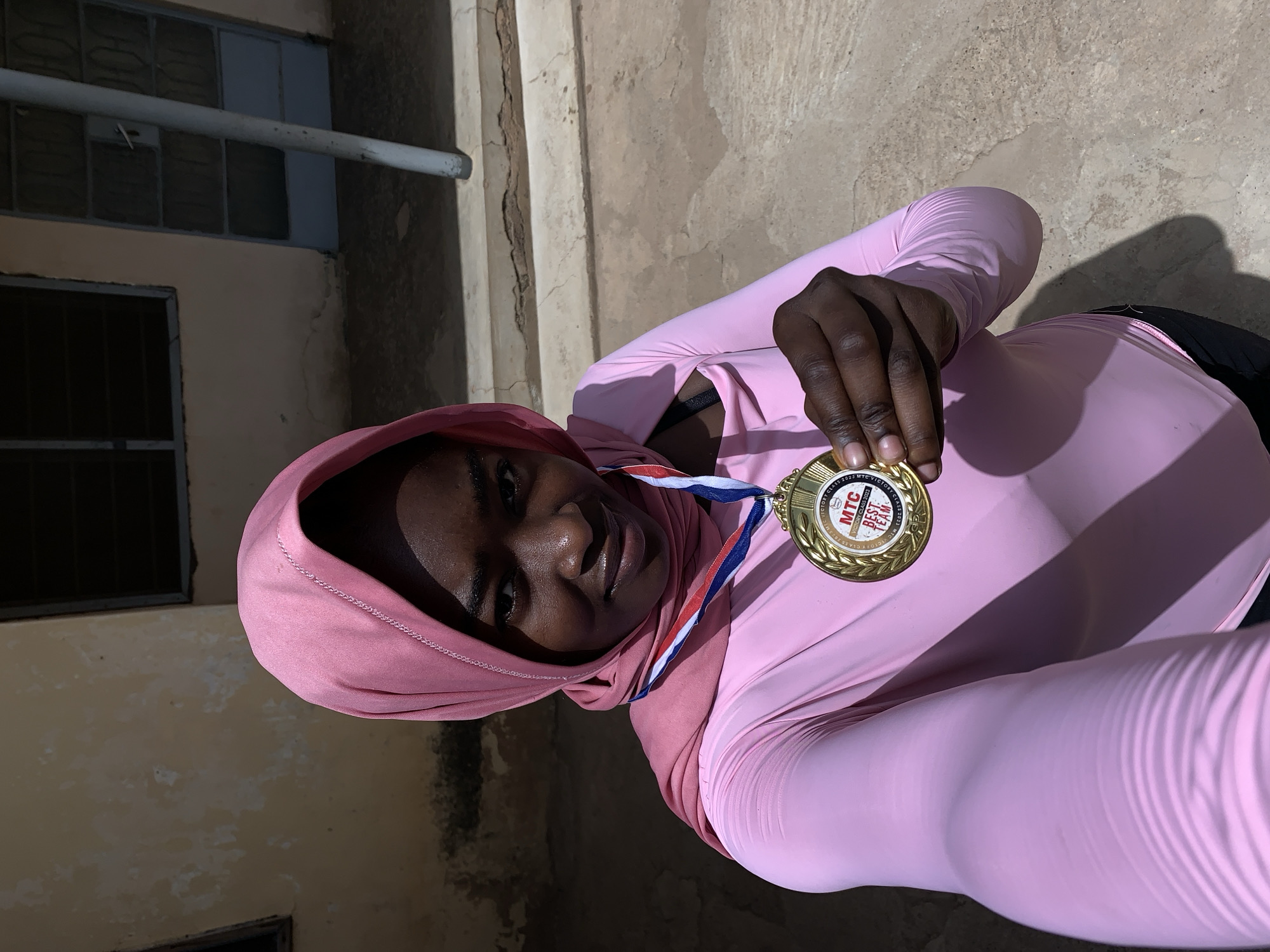 A young woman wearing a pink headscarf and matching long-sleeved shirt is holding a gold medal. The medal is hanging from a red, white, and blue ribbon around her neck. The medal has an inscription that reads "VICTORY CLASS 2023 MTC VICTORY CLASS BEST TEAM." She is smiling slightly and looking at the camera. The background shows a building with a window and a pipe running along the wall. The photograph appears to have been taken in Rafin Par, Nigeria.
