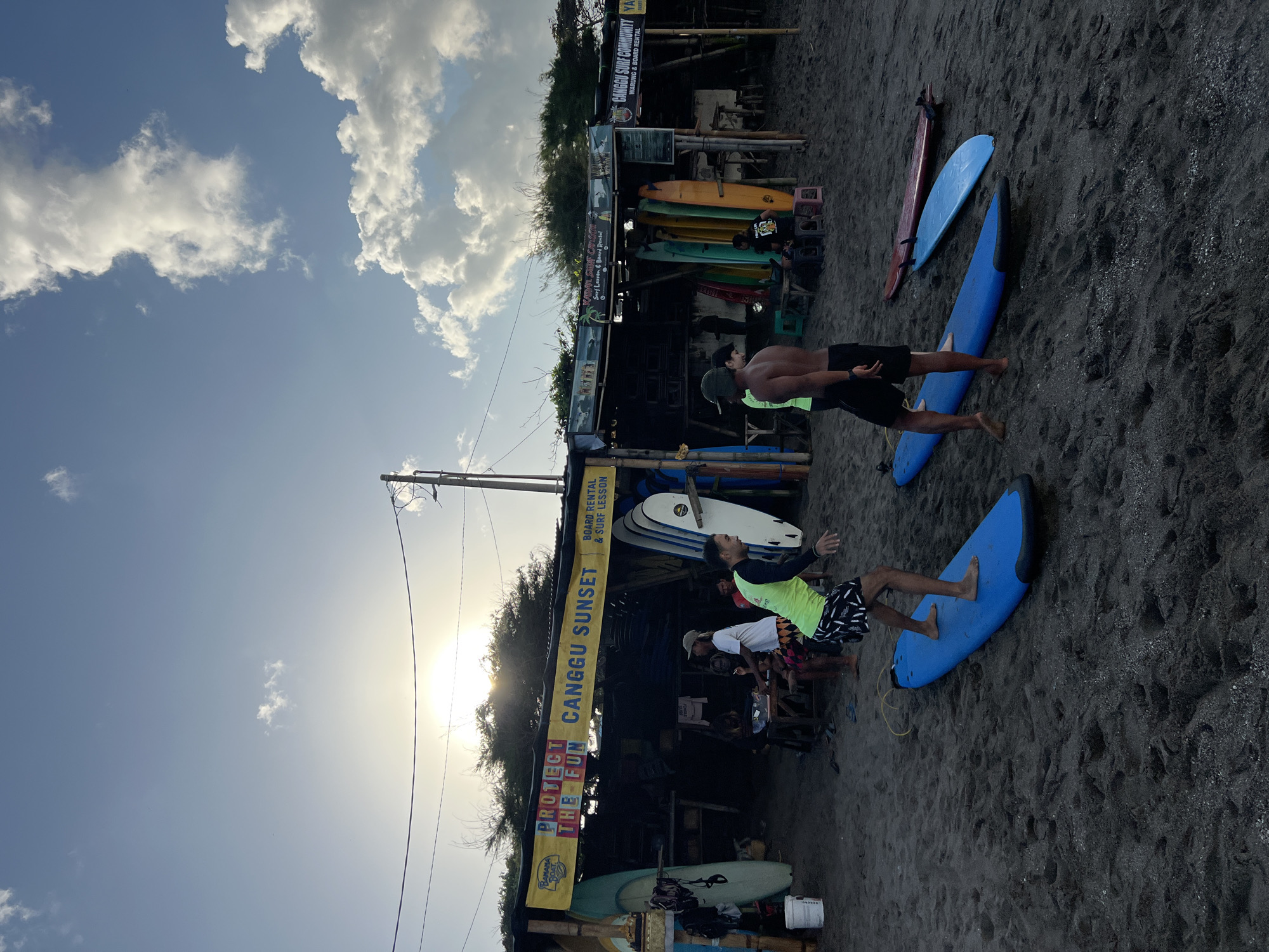 A low-angle, vertical, full outdoor shot captures a surf school on a beach in North Kuta, Indonesia, during dusk. The sky is partly cloudy with the sun peeking through, casting a bright glare. In the foreground, two young men are standing on blue surfboards on dark sand. One man, in a neon yellow rash guard and black and white patterned shorts, is in motion, looking to his right. The other man, shirtless with a dark green cap, is also facing to his right. In the background, a long, open-air structure with a yellow banner that reads "PROTECT THE FUN CANGGU SUNSET BOARD RENTAL & SURF LESSON" houses surfboards of various colors leaning against it. Several people are visible sitting and standing under the structure. The beach is composed of dark, fine-grained sand with footprints visible. The surfboards are arranged upright and lying on the sand, indicating an active surf rental and lesson establishment.
