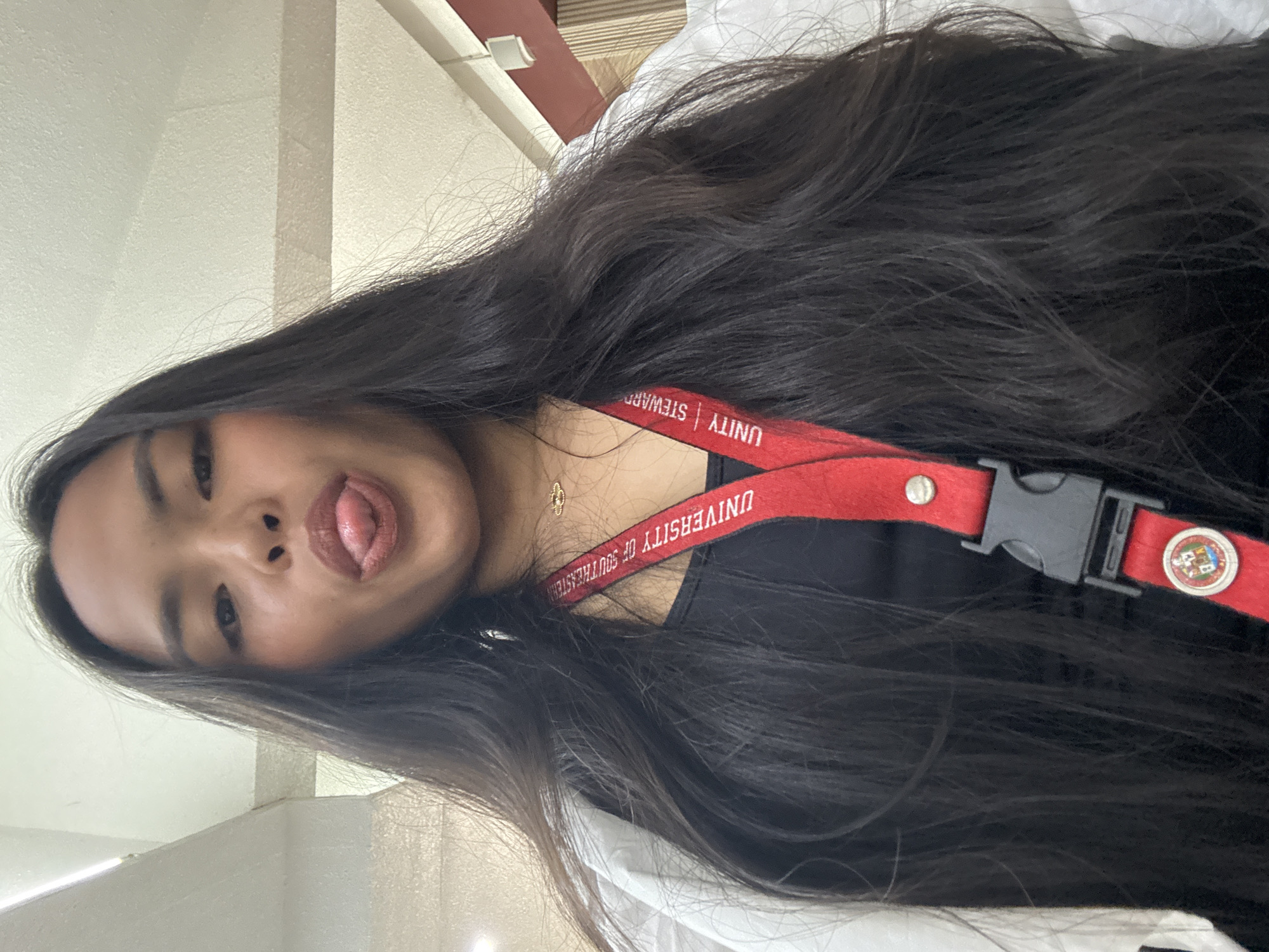 A young woman with long, dark hair and fair skin is posing for a selfie. She is wearing a black top and a white jacket. A red lanyard with the words "UNITY STEWARD" and "UNIVERSITY OF SOUTHEASTERN" printed on it is around her neck. Her tongue is sticking out. The background appears to be an indoor environment with a textured ceiling and a wall with vertical wooden panels. It is not possible to determine the specific location within Davao City from the provided image.