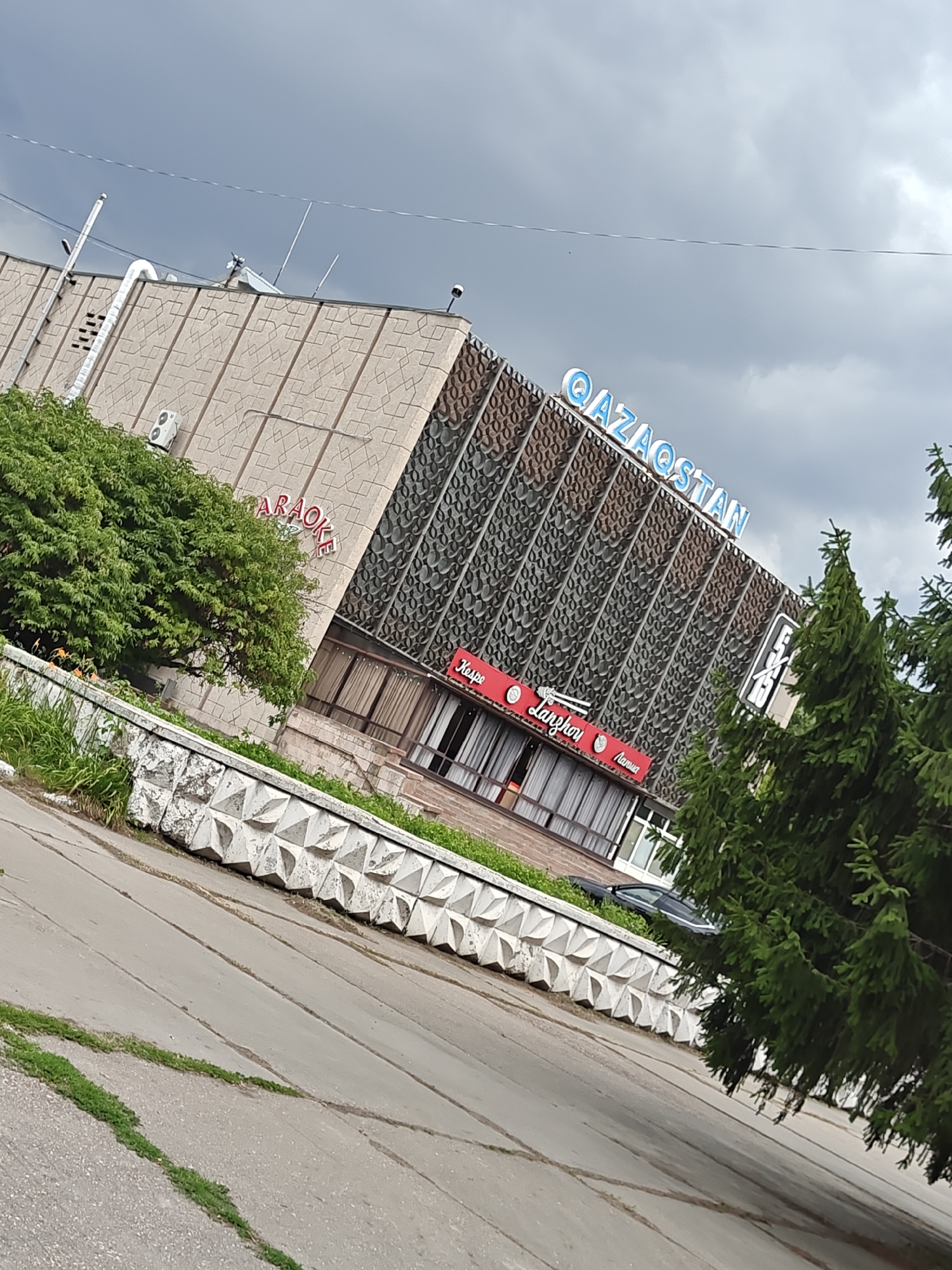 The image depicts the "Qazaqstan" building, a prominent landmark in Petropavl, Kazakhstan. The building features a distinctive facade with a patterned metal screen and a large blue sign displaying the name "QAZAQSTAN". Red signs with Cyrillic text, indicating "Karaoke" and "Lanzhuri", are visible on the lower part of the building. The scene is set against a cloudy sky. In the foreground, a concrete barrier with a geometric design runs along a paved area, likely a sidewalk or road. Lush green trees and bushes surround the building and line the barrier, adding to the urban landscape. A black car is partially visible parked behind the concrete barrier. The perspective is from a slightly elevated angle, looking up at the building.