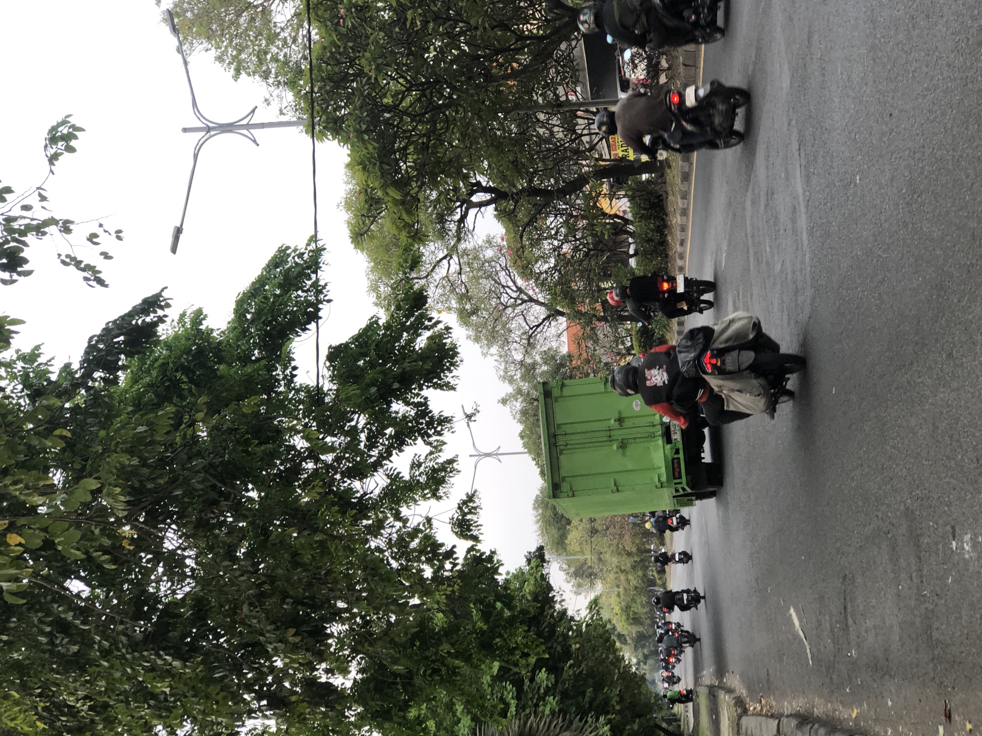 A convoy of motorcycles, some with passengers, travels along a paved road lined with trees and vegetation. A large, green truck is positioned in the foreground, partially obscuring the view of the motorcycles behind it. The sky is overcast. The scene appears to be a typical street in Sumorame, Indonesia, characterized by its lush greenery and busy road traffic, common in urban and semi-urban areas of the country. The motorcycles are likely a common mode of transportation in this region.