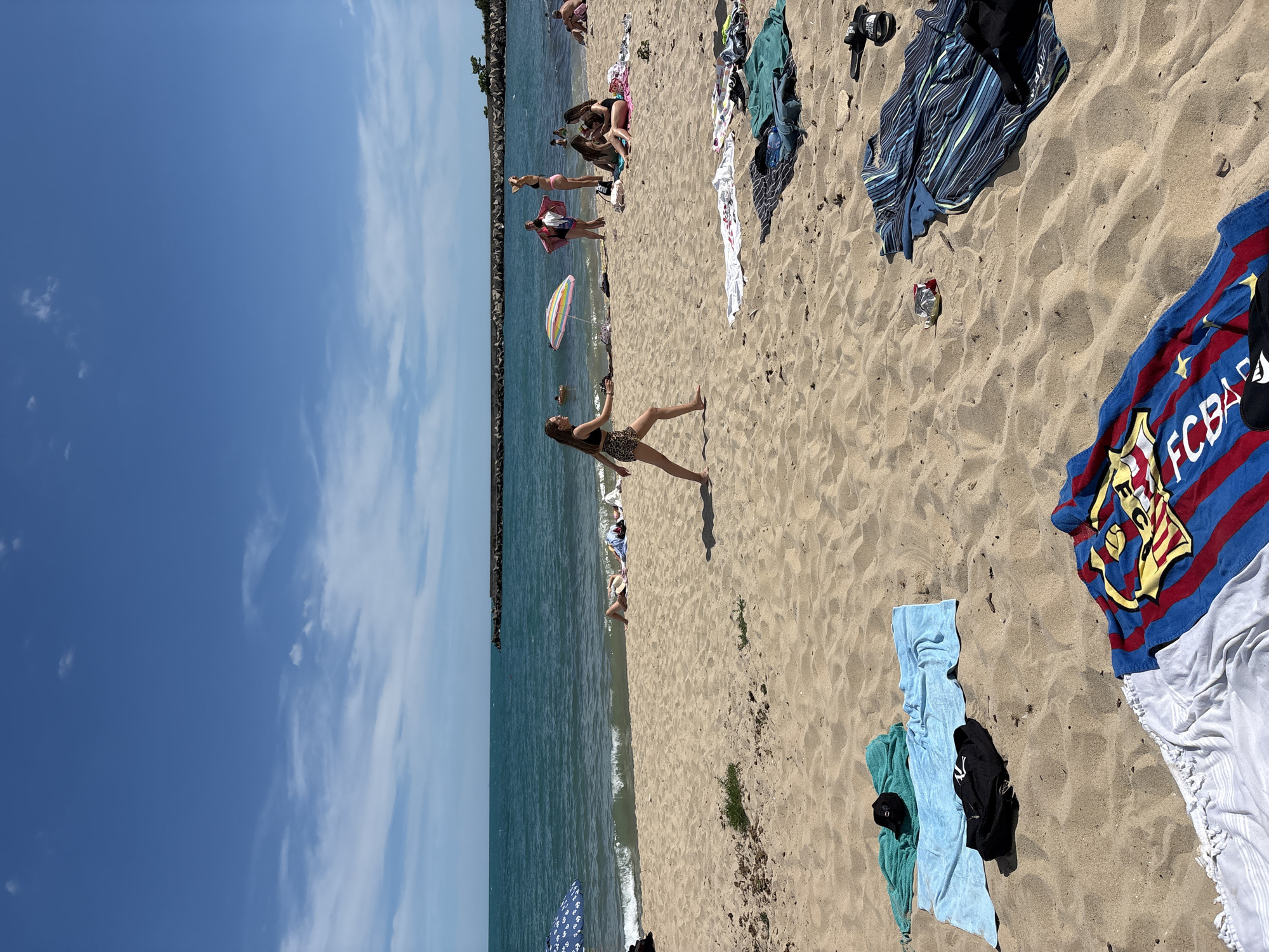 On a sunny day at the beach in Varna, Bulgaria, people are enjoying the sand and sea. Several individuals are scattered across the sandy shore, some lying on towels and others sitting. In the foreground, towels of various colors are laid out on the sand. A young woman in a black bikini top and leopard-print shorts is walking across the beach. In the mid-ground, several other people are gathered, some in swimwear. An umbrella with colorful stripes provides shade. The sea is a vibrant turquoise, with gentle waves lapping at the shore. A stone breakwater extends into the sea in the distance. The sky is a clear blue with a few scattered white clouds.