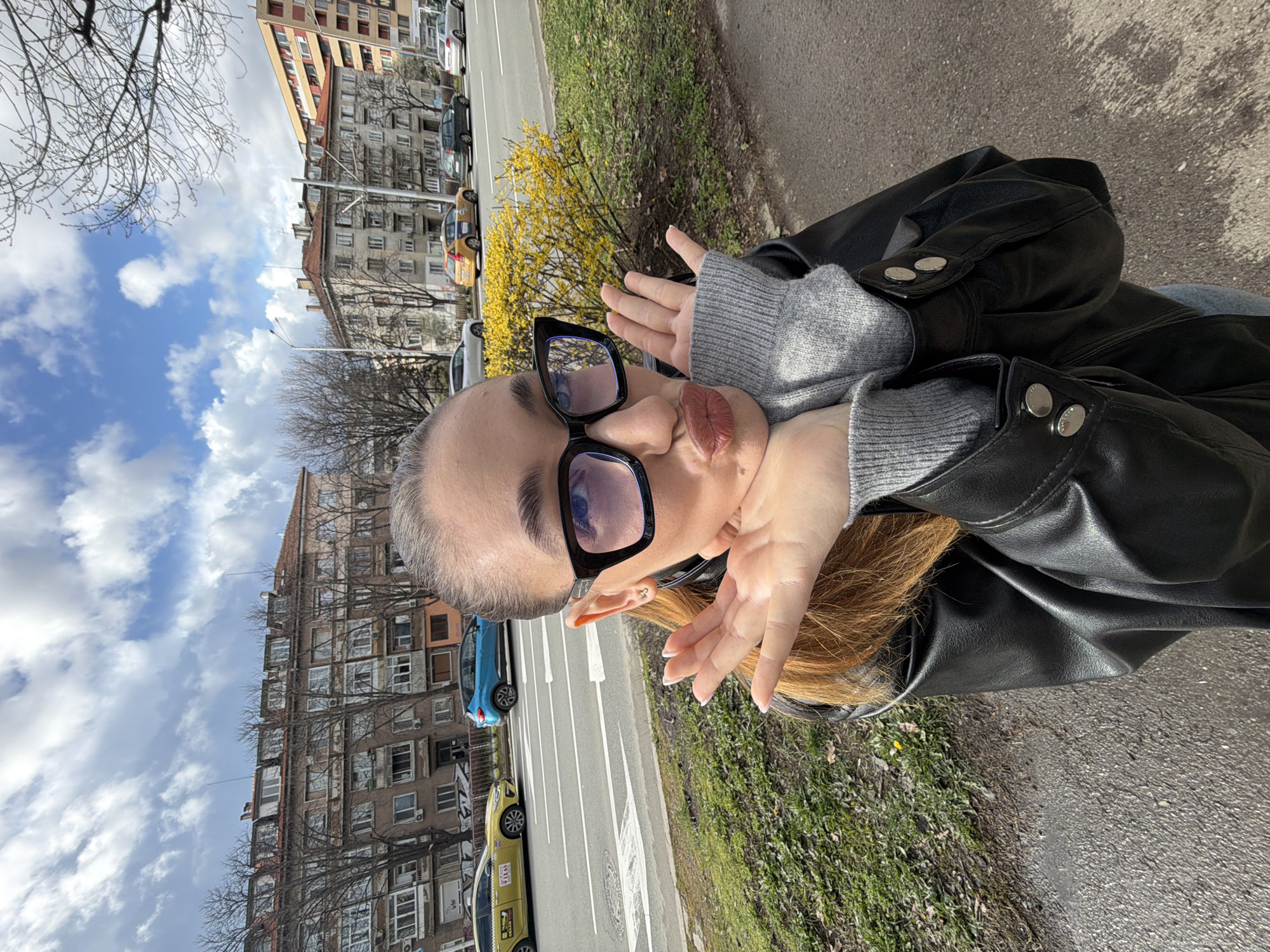 A woman with gray hair, wearing large black-framed glasses, a gray sweater, and a black leather jacket, is posing for a selfie on a street. The location appears to be in Sofia, Bulgaria, based on the architecture visible in the background. Behind her, there is a multi-lane road with several parked cars, including a yellow taxi and a blue car. A row of buildings line the opposite side of the street, exhibiting a mix of residential and commercial use. The sky is partly cloudy with blue patches. The immediate foreground consists of the sidewalk and a patch of greenery. A flowering bush with yellow flowers is also visible near the edge of the sidewalk. She is making a gesture with her hands framing her face.