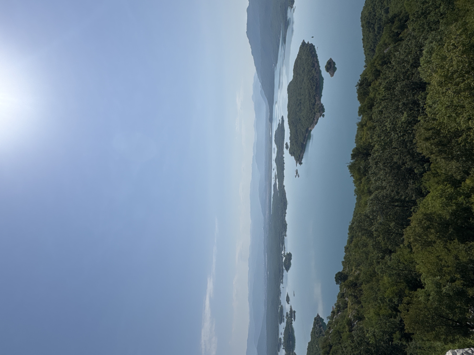 The image captures a vast, serene lake dotted with numerous green, tree-covered islands. The water is a tranquil blue, reflecting the clear, bright sky overhead. In the foreground, a dense forest of lush green trees covers a hillside. In the distance, a series of hazy blue mountains form a majestic backdrop. The sun is high in the sky, casting a bright, ethereal glow. The scene depicts the natural beauty of the city of Ponikvica, Montenegro, offering a panoramic view of its picturesque landscape. There are no people, actions, or interactions visible in the image; it is purely a scenic representation of the environment.