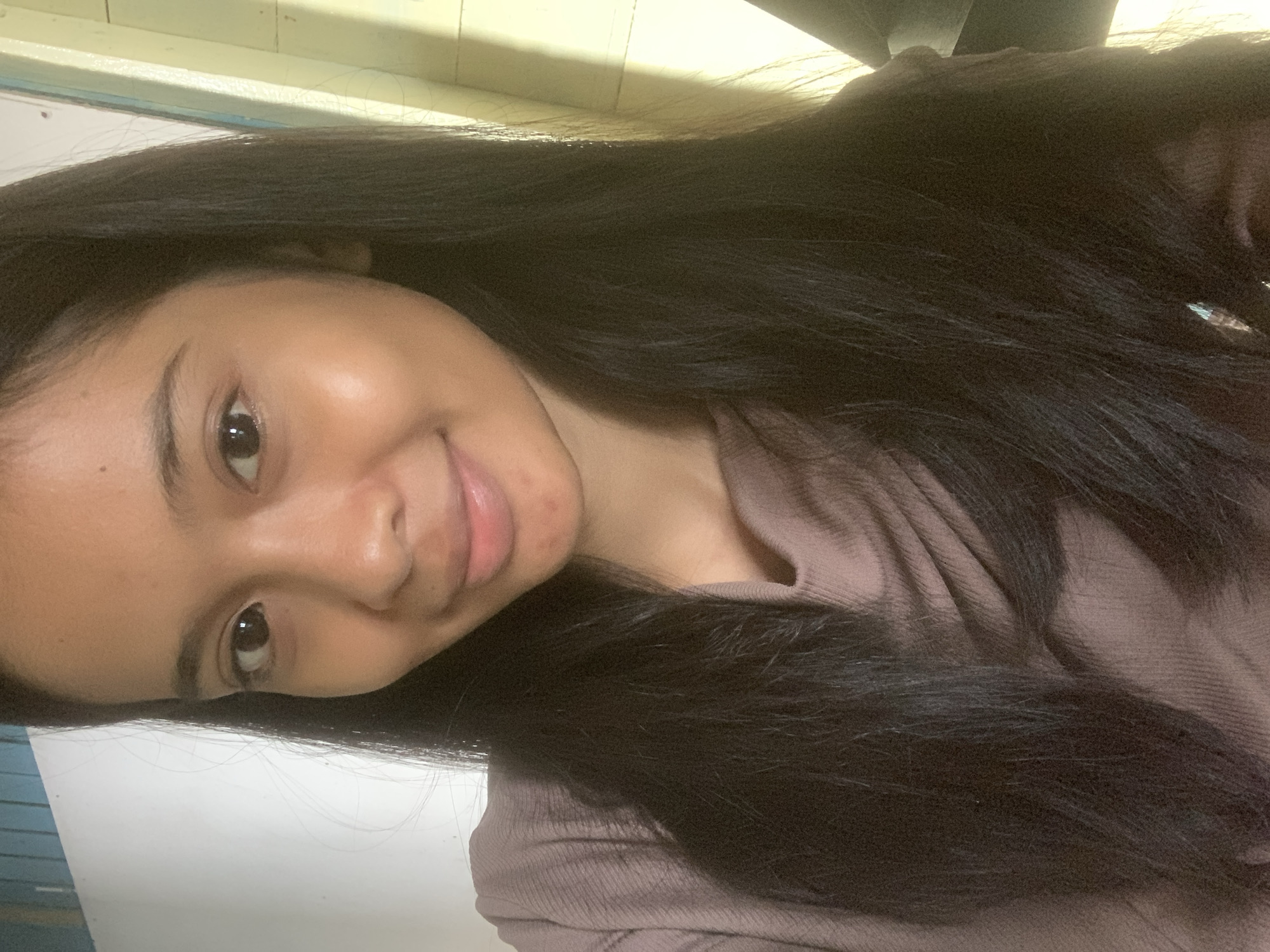 The image is a close-up, waist-up selfie of a young woman with long, dark hair. She is wearing a mauve, cowl-neck top. Her skin appears smooth with a natural glow. Her eyes are dark and she is looking slightly off-camera, with a faint smile. The background is out of focus, but appears to be an indoor setting with a white wall to the left and yellowed wooden paneling to the right, suggesting an interior space possibly in Sungai Meriam, Indonesia. The lighting creates some shadows and highlights on her face and hair. There are no other people or significant objects visible in the frame.