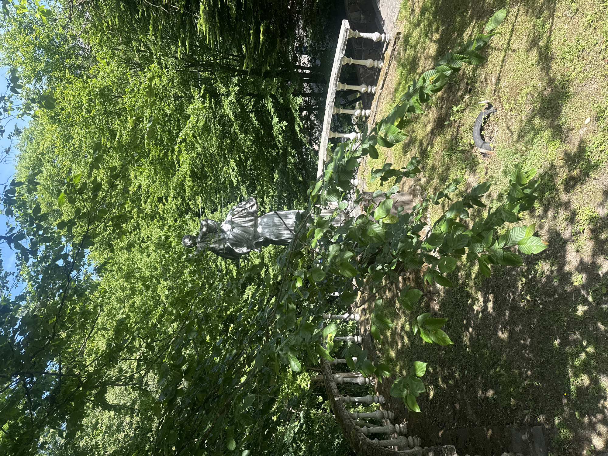 The image depicts a statue of a woman in an outdoor setting. The statue stands on a grassy area near a stone balustrade. The background is filled with dense green trees. A discarded tire is also visible in the grassy area. The location is Syniak, Ukraine.