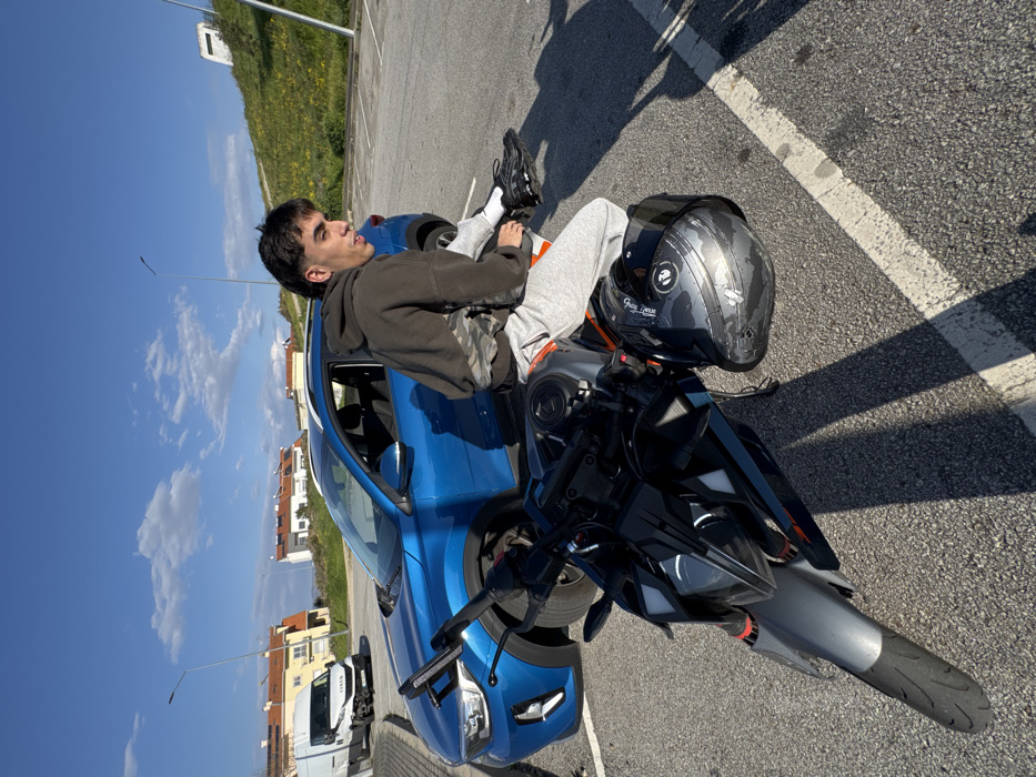 The image depicts an outdoor scene in Mirandela, Portugal, set within a paved parking area under a clear blue sky with scattered cumulus clouds. In the foreground, a male individual with dark hair styled in a mullet sits on a grey, black, and orange sport-naked motorcycle. He is wearing a dark hooded sweatshirt, light grey sweatpants, and black athletic shoes with white socks, looking slightly to his left. A scuffed black full-face motorcycle helmet rests on the motorcycle's tank or seat area. Behind the motorcycle, a blue compact crossover SUV, consistent with a Kia Stonic model, is parked. Further in the background, a white Iveco commercial van is visible on an adjacent road, its front passenger-side bumper appearing damaged or dislodged. Residential buildings with light-colored facades, red-tiled roofs, and visible solar panels are in the mid-ground, while a grassy, unkempt slope rises on the right side of the background. Streetlight poles are present against the sky.