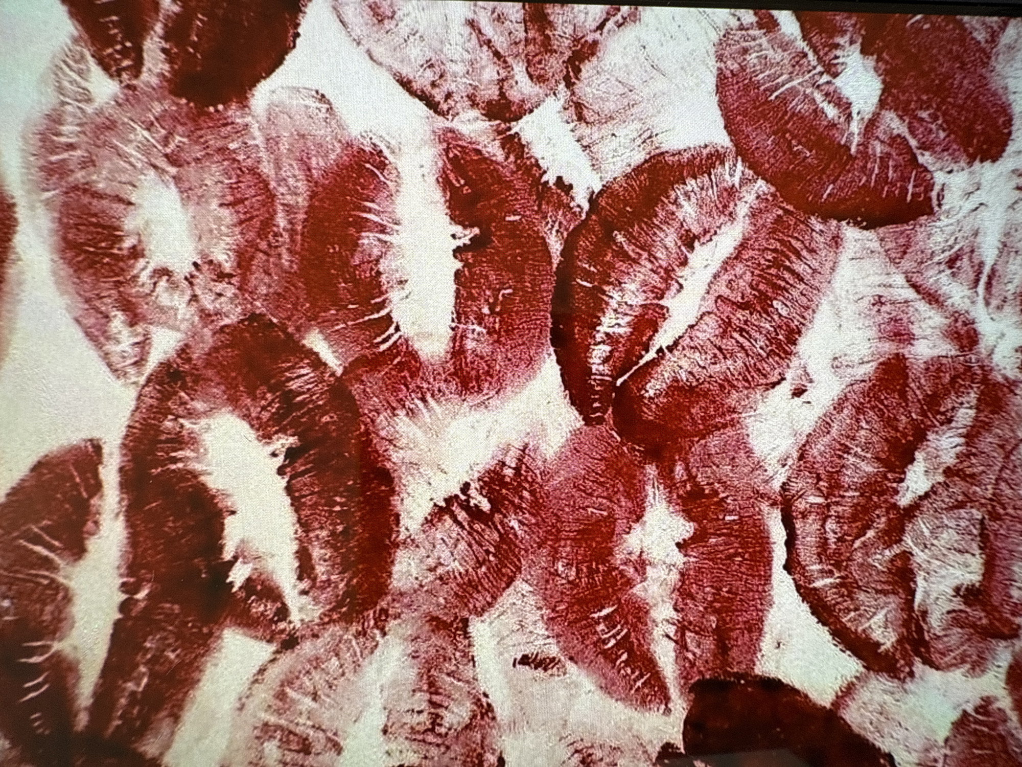 The image presents a dense arrangement of numerous red lipstick kiss prints covering a white, subtly textured surface. The prints exhibit varying intensities of a deep, vivid red, with some displaying clear labial lines and contours, while others appear slightly smudged or faded. The individual imprints frequently overlap, creating a layered effect across the entire field of view. No specific individuals are present in the frame; however, the presence of the prints signifies the repeated action of human lips pressing against the surface. This visual detail is situated within a location in the city of Budva, Montenegro.