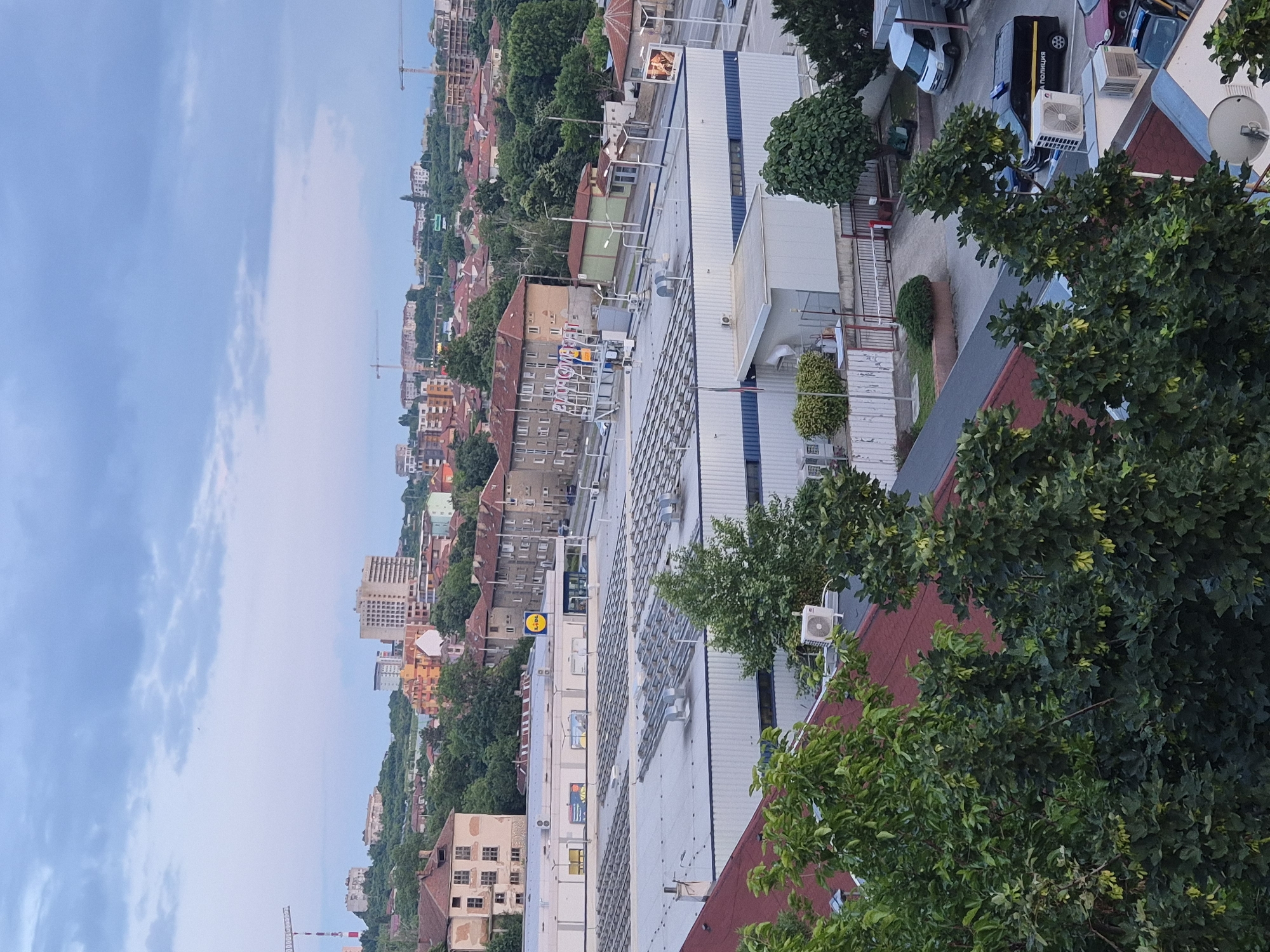 An elevated view captures an urban and suburban landscape under an overcast or twilight sky within an unknown city. In the immediate foreground, dark green leafy trees with light green seed pods partially obscure red and dark grey rooftops equipped with white air conditioning units and a single satellite dish.
The midground features a prominent, large white commercial building with a dark blue stripe near its top, presenting a wide, grey roof covered with numerous rectangular rooftop units, likely ventilation systems or solar panels. To the left and slightly behind this building, another larger structure with a light facade displays a yellow Lidl supermarket logo. Further right, a building with a sign featuring Cyrillic characters spelling "НАРОДЕН МАГАЗИН" (People's Store/Supermarket) is visible. Numerous multi-story residential buildings with red-tiled roofs are scattered throughout the midground and background, interspersed with dense green foliage.
The background reveals a hilly terrain with more residential and commercial structures, several active construction cranes indicating ongoing development, and a tall, light-colored high-rise building. A paved parking area is visible between the foreground structures and the larger commercial building.
Vehicles observed include a white commercial van and a black police van with "ПОЛИЦИЯ" (Police) written in white Cyrillic script on its side, parked near the foreground trees and buildings. Several other cars are partially visible. No individuals are discernible within the scene.