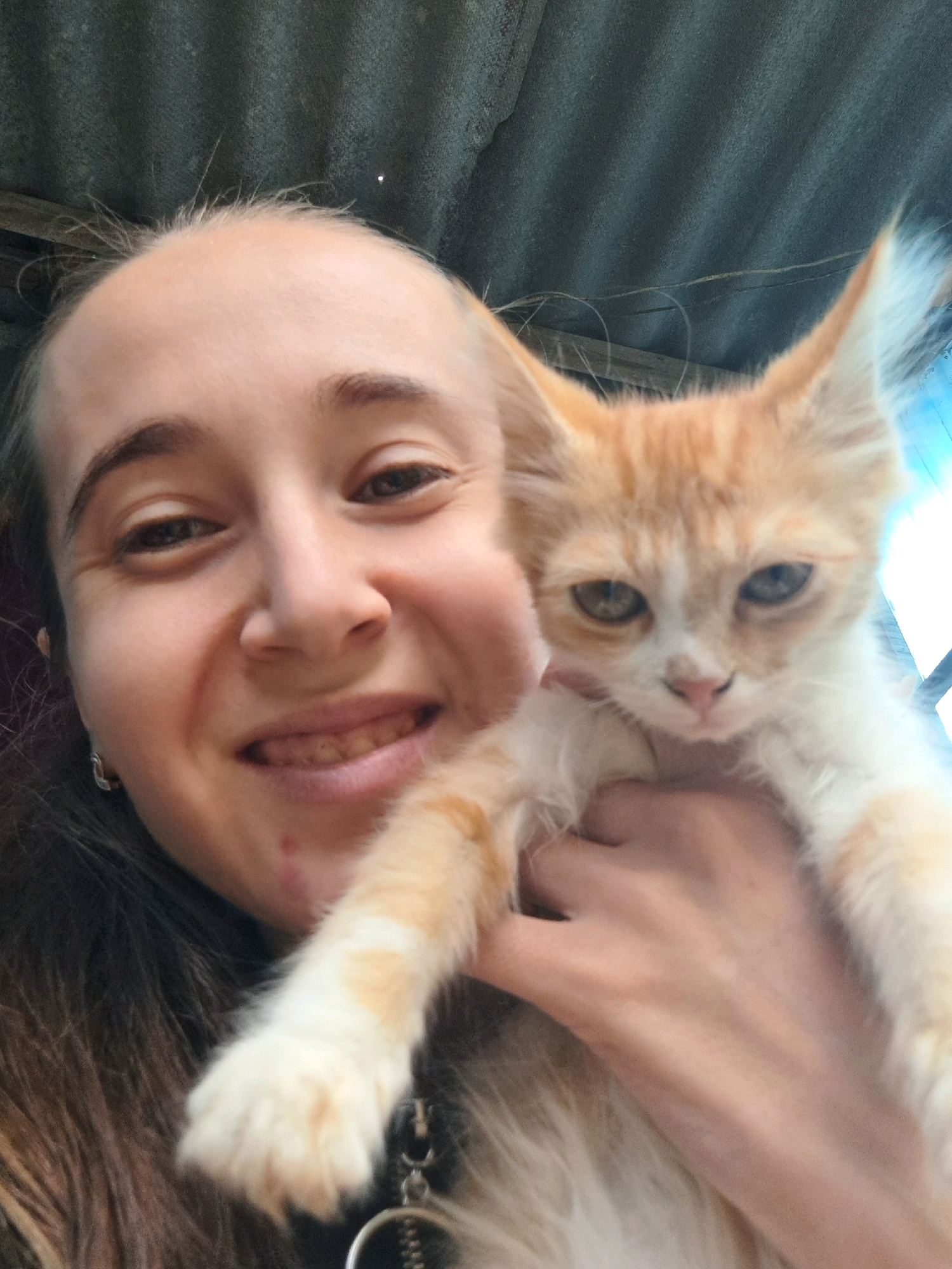A selfie is taken by a young woman holding an orange and white kitten. The woman's head and shoulders are in the foreground, with the kitten held in her right arm. She is smiling broadly, with her teeth visible. The kitten's face is turned towards the camera, with its ears pointed up. The background is a corrugated metal roof. The image appears to be taken outdoors, possibly in a rural or semi-rural setting. The location is Vypasne, Ukraine.