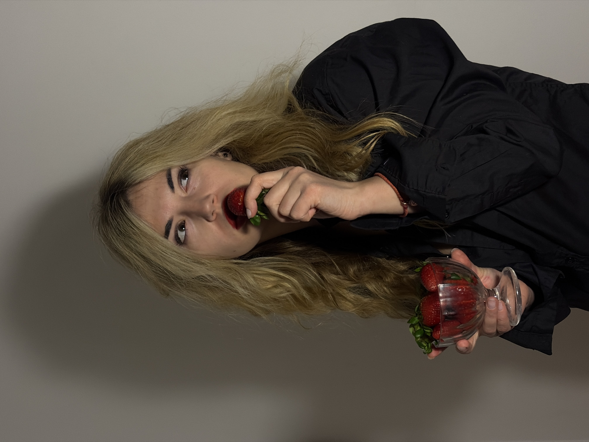 A woman with long blonde hair, wearing a black jacket, is biting into a strawberry. She is holding a glass filled with more strawberries in her other hand. The background is a plain light grey wall. The image does not show any information that identifies the location as Budva, Montenegro.