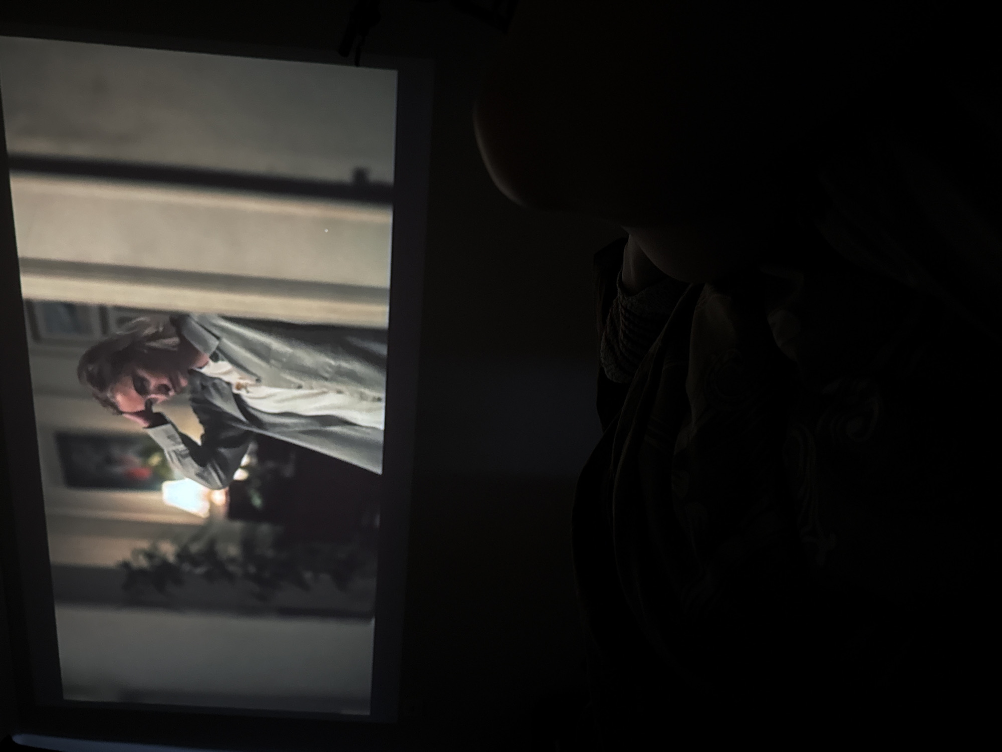 The image displays a dark room with a brightly lit projection screen on the left, contrasting with the nearly black right side where a person is dimly visible.
On the projector screen, a video frame is shown, featuring a person, likely male, with light-colored hair. He is wearing a light-colored shirt or blouse underneath a darker jacket or coat. His gaze is directed downwards and to the left within the frame, and his right hand is raised towards his face, though it's unclear if he's holding an object. The projected image is rotated 90 degrees counter-clockwise. The background of the projected scene appears to be an indoor setting, with a visible light source (possibly a lamp or wall fixture), blurry elements that could be furniture or plants, and horizontal architectural details in the upper section. The lighting within the projected scene is moderate, suggesting an indoor setting illuminated by artificial light.
To the right, a person is partially visible in silhouette against the dark background. Only the outline of their head, neck, and shoulder, along with a portion of patterned clothing (possibly a sweater), can be faintly distinguished. This individual appears to be an observer, facing towards the projected screen. The surrounding room is profoundly dark, indicating either nighttime or that the environment has been darkened for optimal projector viewing. No specific objects are discernible in the dark portion of the room, and there is no visible text in the entire image.
