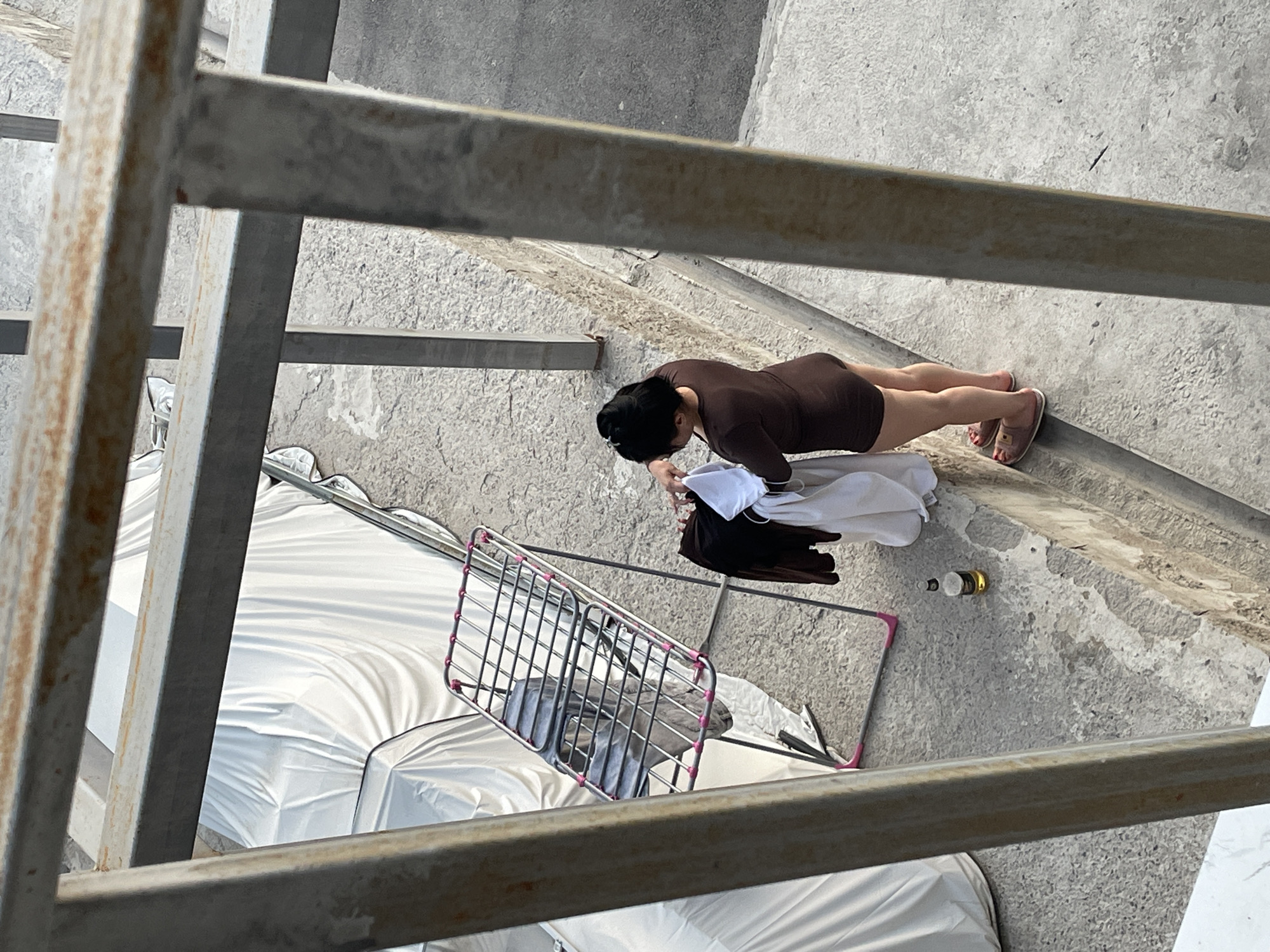 The image shows a woman in a brown outfit standing next to a drying rack with clothes. She appears to be folding or arranging laundry. A bottle of what looks like beer is on the ground nearby. The scene is outdoors, with a concrete ground and a wall. In the background, there are vehicles covered with white tarps. The overall setting suggests a residential or possibly a storage area in Almaty, Kazakhstan. The image is framed by rusty metal bars, indicating it was taken from a higher vantage point, possibly a balcony or window.