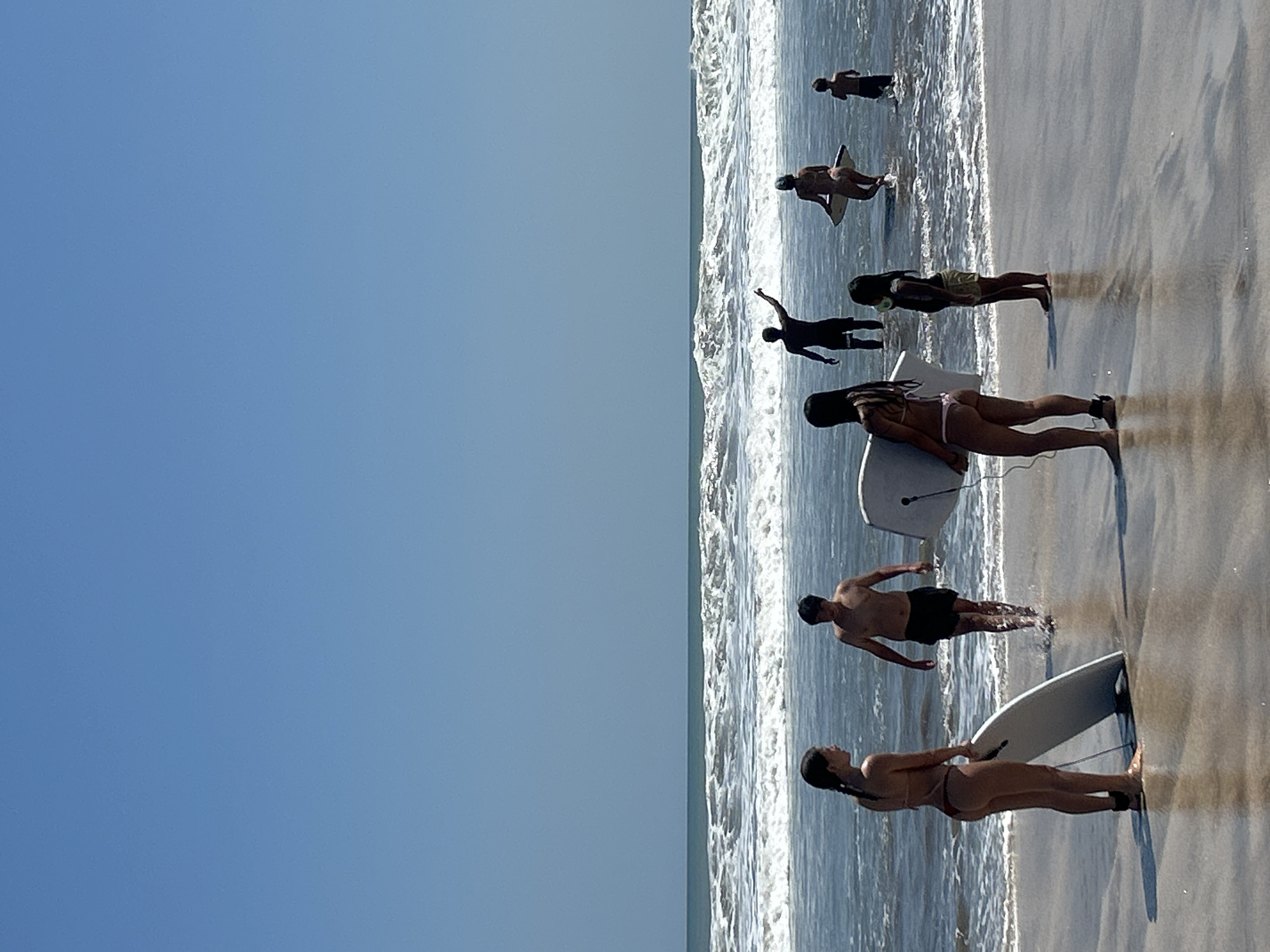 The image depicts a beach scene in Kuta, Indonesia, on a clear, sunny day with a vast blue sky. The ocean is active with moderate waves breaking near the shore. Several individuals are present on the wet sand and in the shallow water.
In the foreground, on the left, a young woman wearing a bikini stands with her back to the camera, holding a white boogie board. To her right, a young man in dark shorts walks through the shallow water, also with his back partially turned. Further to the right and slightly behind them, another young woman, also in a bikini, is seen from the back, holding a white boogie board. To her right, a young child in shorts stands in the water.
In the mid-ground, a group of people are in the water. A man in dark shorts is visible in the middle, with one arm raised and pointing towards the ocean, seemingly instructing or gesturing. To his right, a person in a bikini is walking in the water with a boogie board. Further to the right, a young child is in the water. In the background, several other figures are visible further out in the ocean, some appearing to be in deeper water.
The sand appears wet and reflective from the incoming tide. The overall scene captures a group of people enjoying recreational activities at the beach.