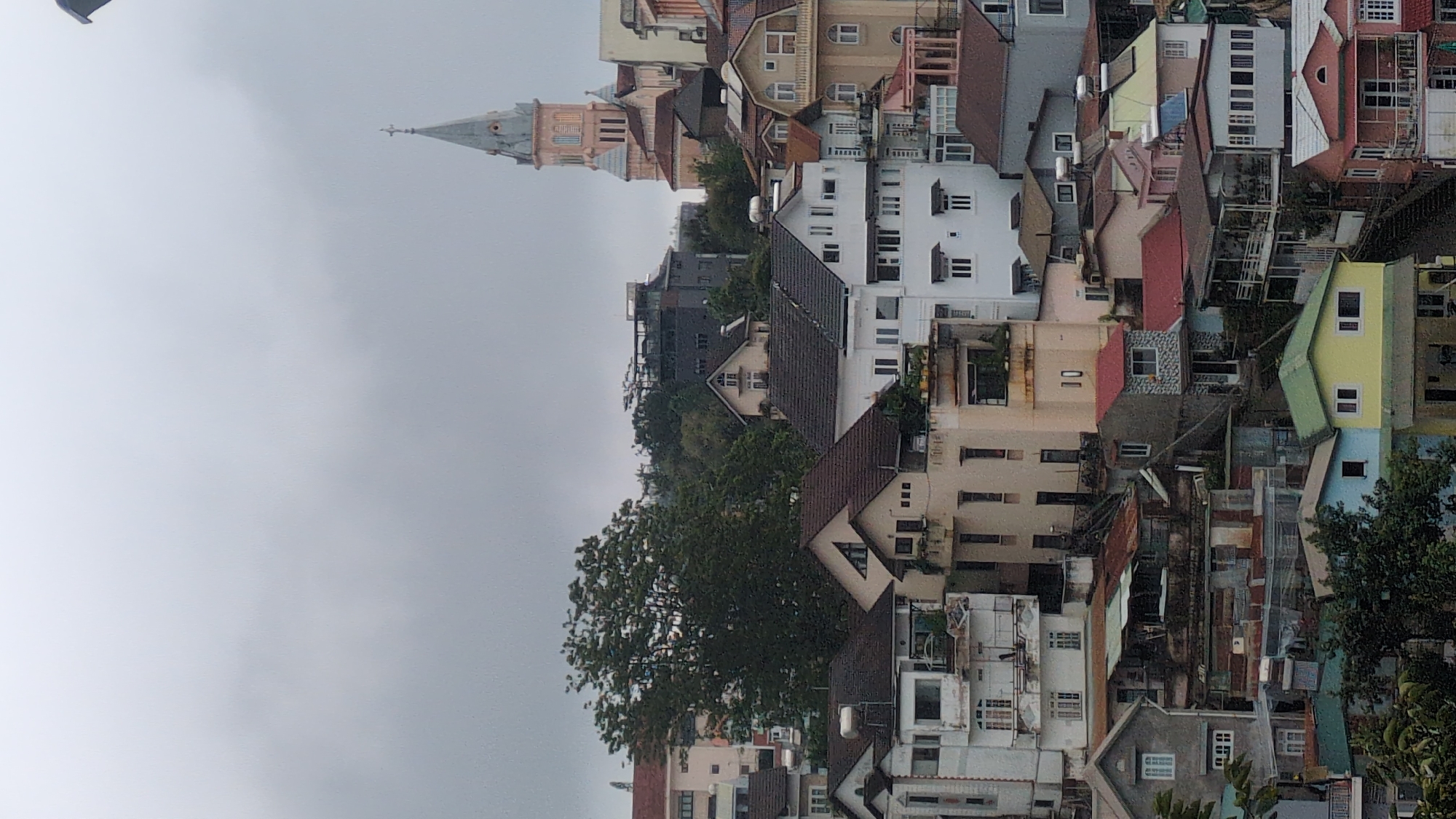 This is a medium-distance, eye-level, vertical shot of a dense urban hillside neighborhood in Da Lat, Vietnam, under an overcast sky. The scene is dominated by a cluster of houses and buildings of various sizes and architectural styles, stacked closely together on a steep incline. The buildings are a mix of colors, including white, cream, beige, gray, light pink, and some with red roofs. Many of the structures appear to be multi-story homes with balconies and pitched roofs, some covered in dark tiles and others with weathered corrugated metal. A prominent pink and cream colored church with a tall, slender spire stands out in the upper right portion of the frame, overlooking the neighborhood. Dense green trees are scattered throughout the landscape, with a particularly large tree in the mid-left. The overall impression is one of a densely populated, hillside community with a blend of older and more modern architecture, characteristic of Da Lat.