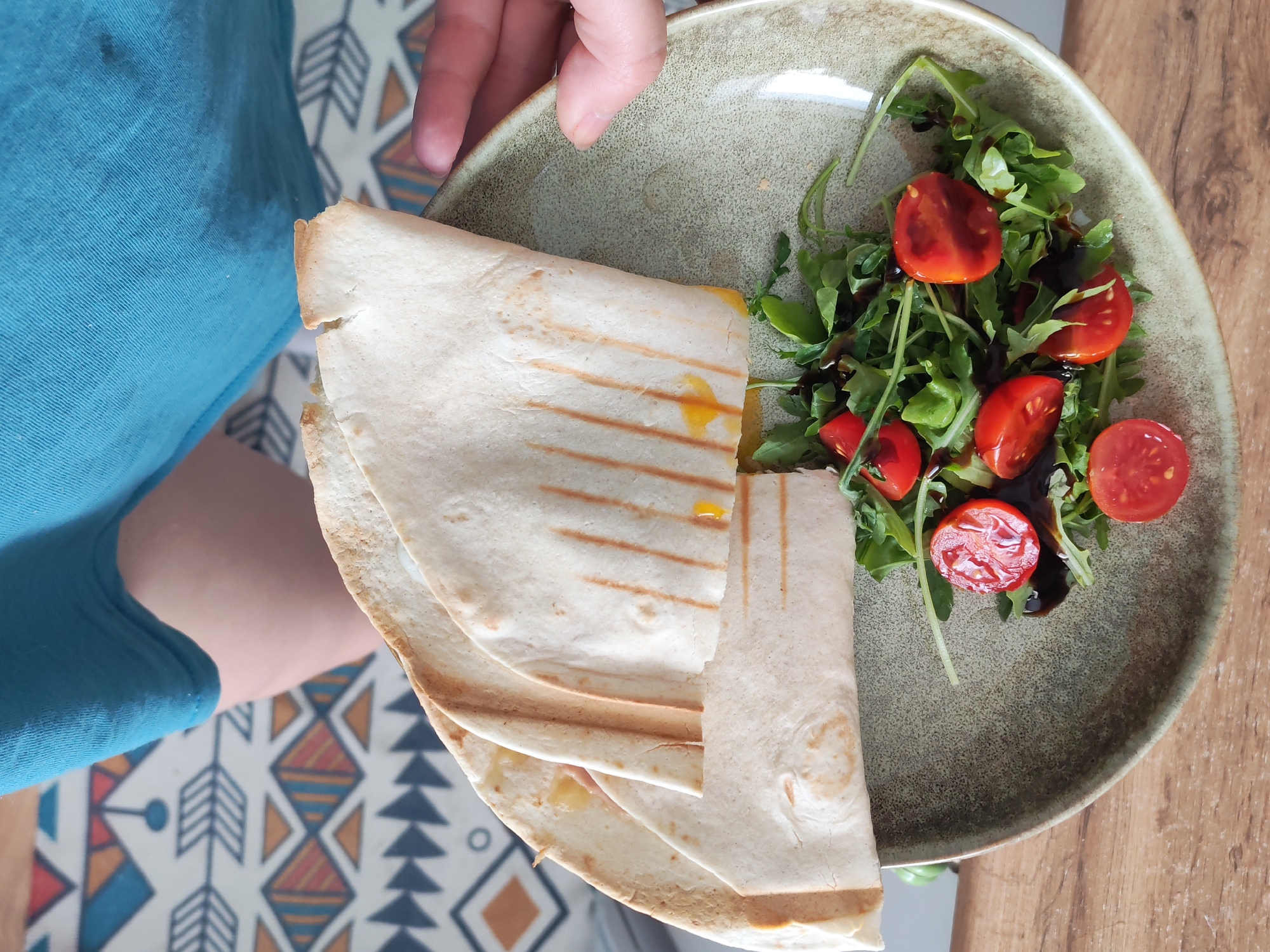 A close-up overhead shot shows a person in a teal shirt holding a plate of food. The plate contains a folded quesadilla with visible grill marks and a side salad of arugula and halved cherry tomatoes, drizzled with a dark sauce. The background includes a patterned placemat and a wooden table. The location is not discernible from the image.