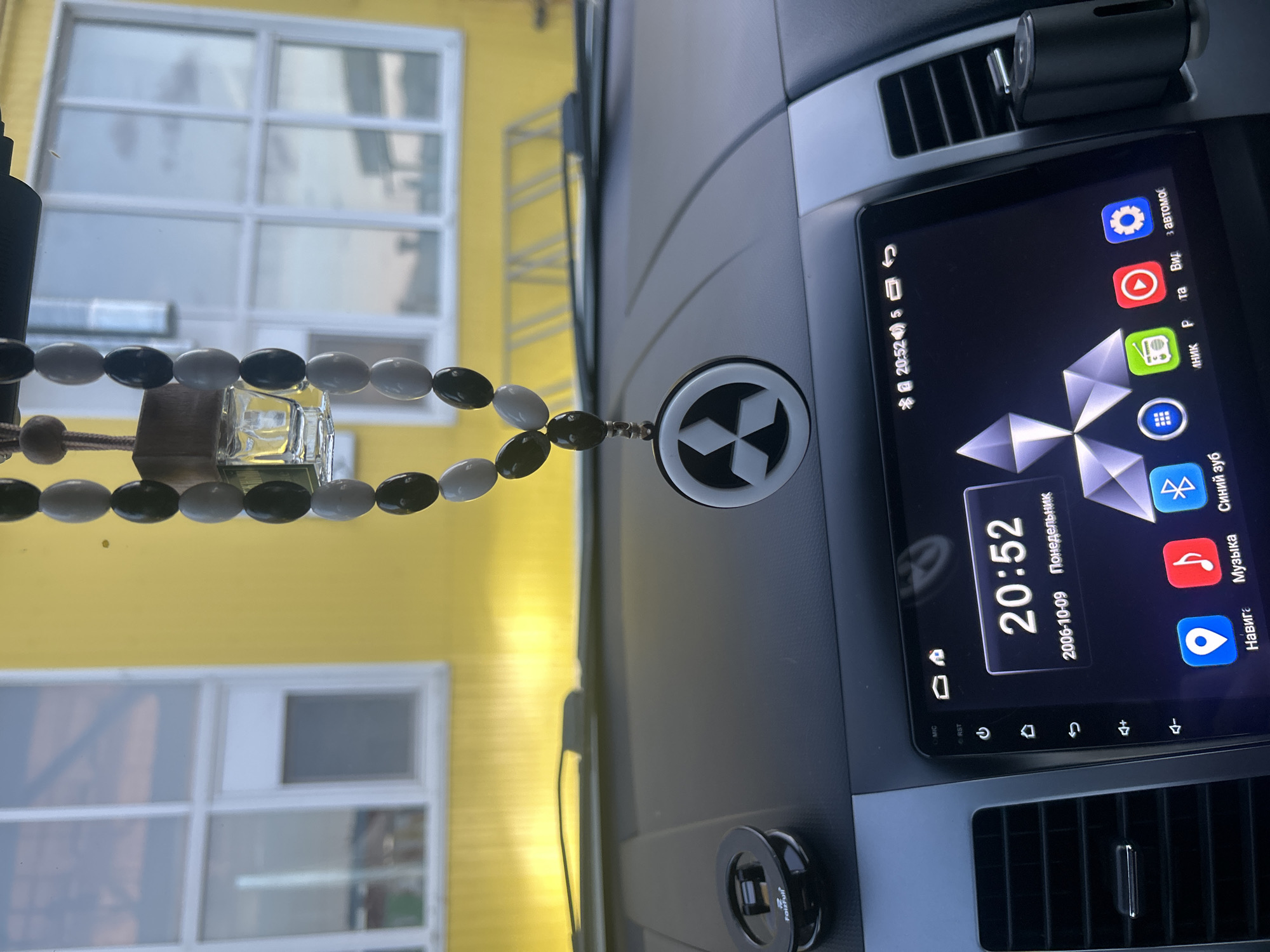 The image depicts the interior of a car. The dashboard is black and houses a digital display showing the time as 20:52 on October 9, 2006, along with various icons and text in Russian. An air freshener, possibly a beaded rosary with a glass vial, hangs from the rearview mirror or dashboard. Through the windshield, a bright yellow building is visible with white-framed windows.
The text "Faizfull" is visible on a device mounted near the bottom of the dashboard.
It's impossible to tell the exact location in Chaikovsky, Russia, based on the visual information.
