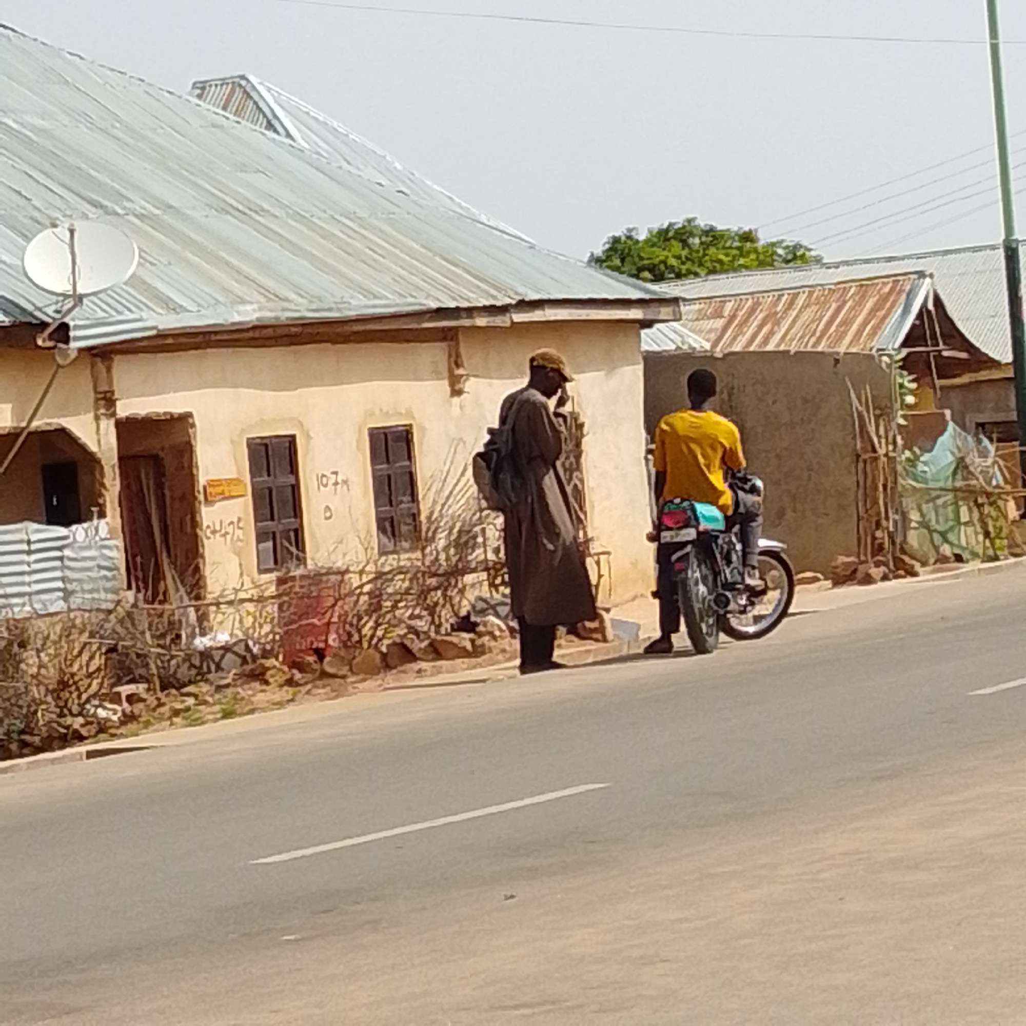 In Gombe, Nigeria, two men are present on a street beside a building. The building has a corrugated metal roof and light-colored walls. The man on the left, wearing a long brown robe and a cap, is standing and appears to be speaking on a phone. He also has a backpack slung over his shoulder. The man on the right is seated on a motorcycle, wearing a yellow t-shirt and dark pants. The motorcycle is facing away from the viewer. The street is paved with a white line marking a lane. The environment appears to be a semi-urban setting with some vegetation visible.