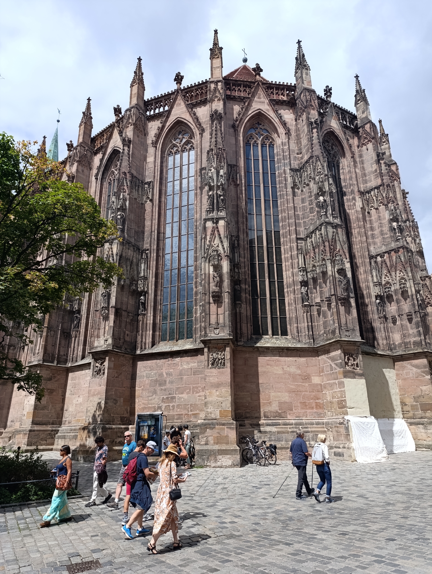 The image depicts a view of the St. Lorenz Church in Nuremberg, Germany, with a clear sky overhead and a cobblestone plaza in the foreground. The towering gothic facade of the church, constructed from reddish-brown stone, features intricate architectural details, including tall pointed windows, spires, and sculptures. To the left of the church, a large green tree with some autumn foliage partially obscures the building.
In the plaza, several people are walking. On the left, a woman in a blue top and a light blue skirt walks away from the camera, carrying a brown bag. A man in a patterned shirt and white pants walks behind her. Further to the right, a group of people are more clustered. Two men in blue shirts and a man in a blue t-shirt and shorts are walking together, one with a red backpack. Another person with a straw hat and a patterned dress is walking in the same direction, holding a map. An older couple is walking towards the right, with the man using a walking stick. Two bicycles are parked near the church wall. The scene suggests a sunny day with a light breeze, indicated by the movement of the tree's leaves.