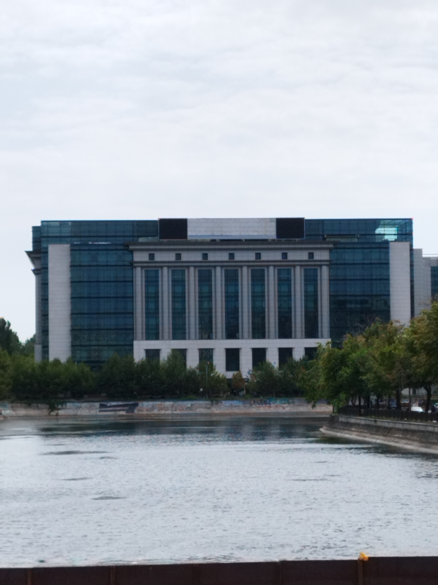 A wide, horizontal shot depicts a large, modern building with a glass facade situated across a body of water. The building features a central section with classical columns and rows of windows, flanked by glass-clad wings. Trees line the bank of the water in front of the building, and the sky above is overcast. The water surface shows subtle ripples, and a dark structure, possibly a barrier or the edge of a walkway, is visible in the foreground. The scene appears to be the National Library of Romania in Bucharest.