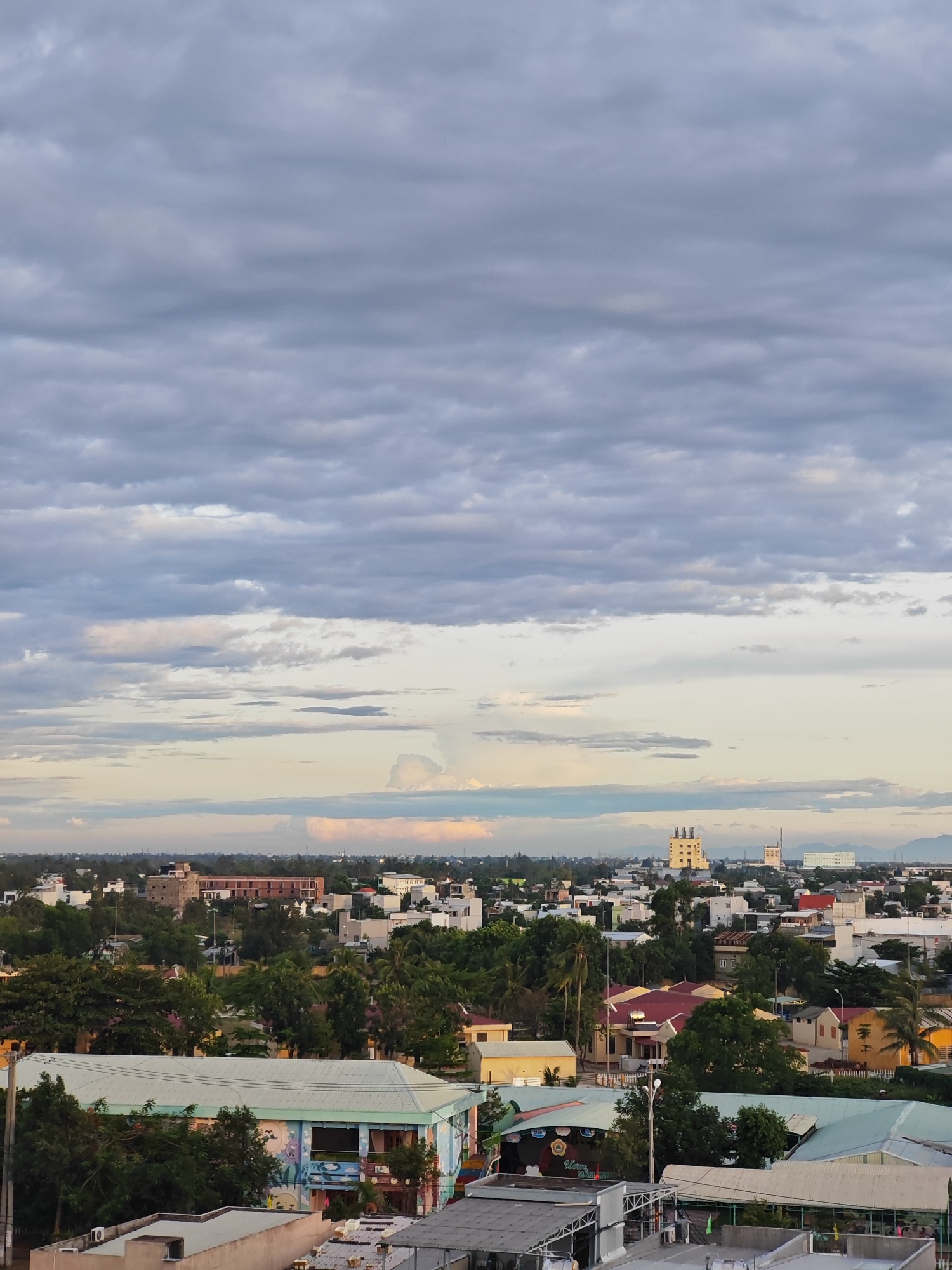 This is a wide-angle, eye-level photograph taken from a high vantage point overlooking a city and its surrounding environment. The scene is dominated by a vast expanse of clouds that take up the upper two-thirds of the frame. These clouds are layered and varied in texture, ranging from dense, dark grey formations in the upper portion to lighter, softer grey and white wisps closer to the horizon. The lighting suggests it is either early morning or late afternoon, with a soft, diffused light filtering through the cloud cover.
Below the clouds, a low-lying cityscape is visible. Buildings of varying sizes and architectural styles are densely packed together. Many of the structures are residential homes with pitched roofs, while there are also some larger, more modern buildings, including what appear to be multi-story residential or commercial blocks. The city is interspersed with abundant greenery, with numerous trees providing a lush canopy over parts of the urban landscape. The overall impression is one of a developed but not overly industrialized urban area, with a significant presence of natural vegetation.
In the foreground, closer buildings are visible, some with distinct features like colorful murals on their facades and metal awnings. The rooftops of these structures show varied construction materials and designs, including corrugated metal and tiled roofs. The presence of air conditioning units and other rooftop equipment suggests an inhabited and functional environment.
The sky transitions from the heavy cloud cover to a softer, lighter sky near the horizon, with hints of warmer tones like pale yellow and pink, possibly indicating the sun's position. In the far distance, faint outlines of mountains or hills can be seen, suggesting the city is situated in a region with some natural topographical features.
While the image provides a broad overview of the city, there are no identifiable people, specific landmarks, or vehicles that would pinpoint an exact location with certainty. However, the architectural style, building density, and prevalence of tropical or subtropical vegetation could be indicative of Southeast Asian cities. Given the information that this is a location in Đà Nẵng, Vietnam, the scene aligns with the characteristic appearance of urban areas in that region. The city appears to be in a period of development with a mix of older and newer constructions, surrounded by significant green spaces.