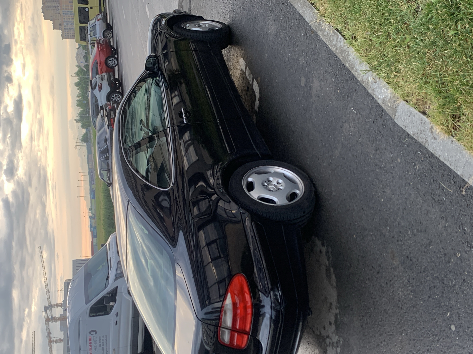 A black Mercedes-Benz CLK coupe is parked on a wet asphalt road in Stavropol, Russia. The car's rear passenger side is prominently featured, showing the taillight, wheel, and part of the door. Reflections of windows and a cloudy sky are visible on the car's glossy surface. In the background, several other cars are parked, including a white van with Cyrillic text, a silver SUV, a red sedan, and a yellow bus. Construction cranes are visible in the distance, suggesting ongoing development. The scene appears to be an outdoor parking area next to a grassy verge, with a concrete curb separating the road from the grass. The sky is overcast with clouds illuminated by the setting sun.