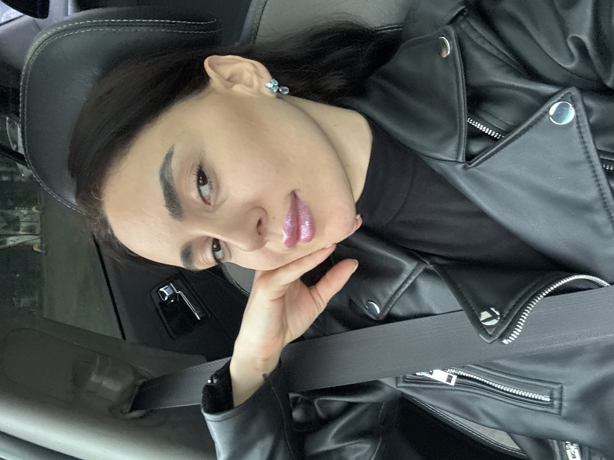 The image depicts a female individual seated in the passenger seat of a vehicle. She has dark hair, dark eyes, and is looking directly at the camera. Her right hand is positioned under her chin/jawline. She is wearing a black faux leather jacket with silver-toned zippers and snap buttons, over a black high-necked top. A black seatbelt is fastened across her chest. A single earring, appearing as a cluster of light blue or white stones, is visible on her right ear. The vehicle's interior features a dark headrest with light stitching, a light grey or beige A-pillar, and a dark door panel with a chrome-colored door handle and lock mechanism. The background visible through the car window is heavily blurred, showing indistinct shapes consistent with an urban street scene, including what appears to be a distant vehicle. The specific location within Tbilisi, Georgia, cannot be identified from the visual information presented in the image.