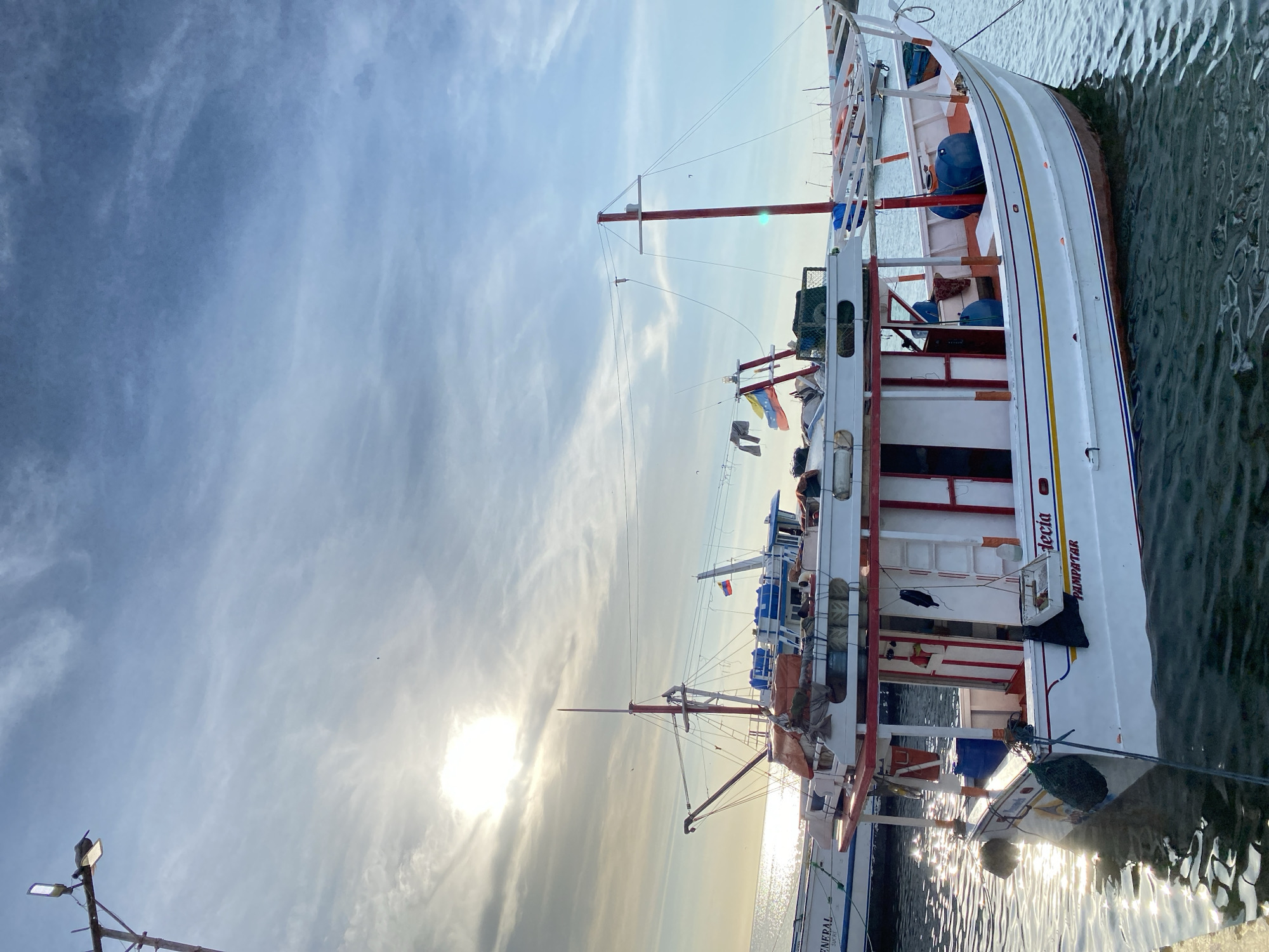The image captures a serene scene at a dock in Juan Griego, Venezuela, featuring a large white fishing boat under a bright, partly cloudy sky. The photograph is oriented vertically, with the boat occupying the right side of the frame, docked in calm water.
The primary subject is a white fishing vessel, possibly a trawler, with a long hull accented by prominent blue and yellow stripes. Red framework, including a tall mast and support beams, provides a striking contrast to the white hull. Various elements of marine life and daily activity are visible on deck: ropes, buoys, fishing nets, and what appear to be clothes hanging out to dry. The boat's name, "Mecia," and "PAMPATAR" are clearly visible in blue text on its stern. A portion of another boat, bearing the text "GENERAL," is visible below the main vessel.
The sun is low in the sky, positioned behind the boats, suggesting either sunrise or sunset. It casts a brilliant glare on the water, creating sparkling reflections that ripple across the surface. The sky is a soft blue with scattered, wispy clouds, indicating clear weather. No people are distinctly visible in the frame, contributing to the tranquil atmosphere. The scene evokes a sense of peaceful inactivity at a fishing harbor or marina.