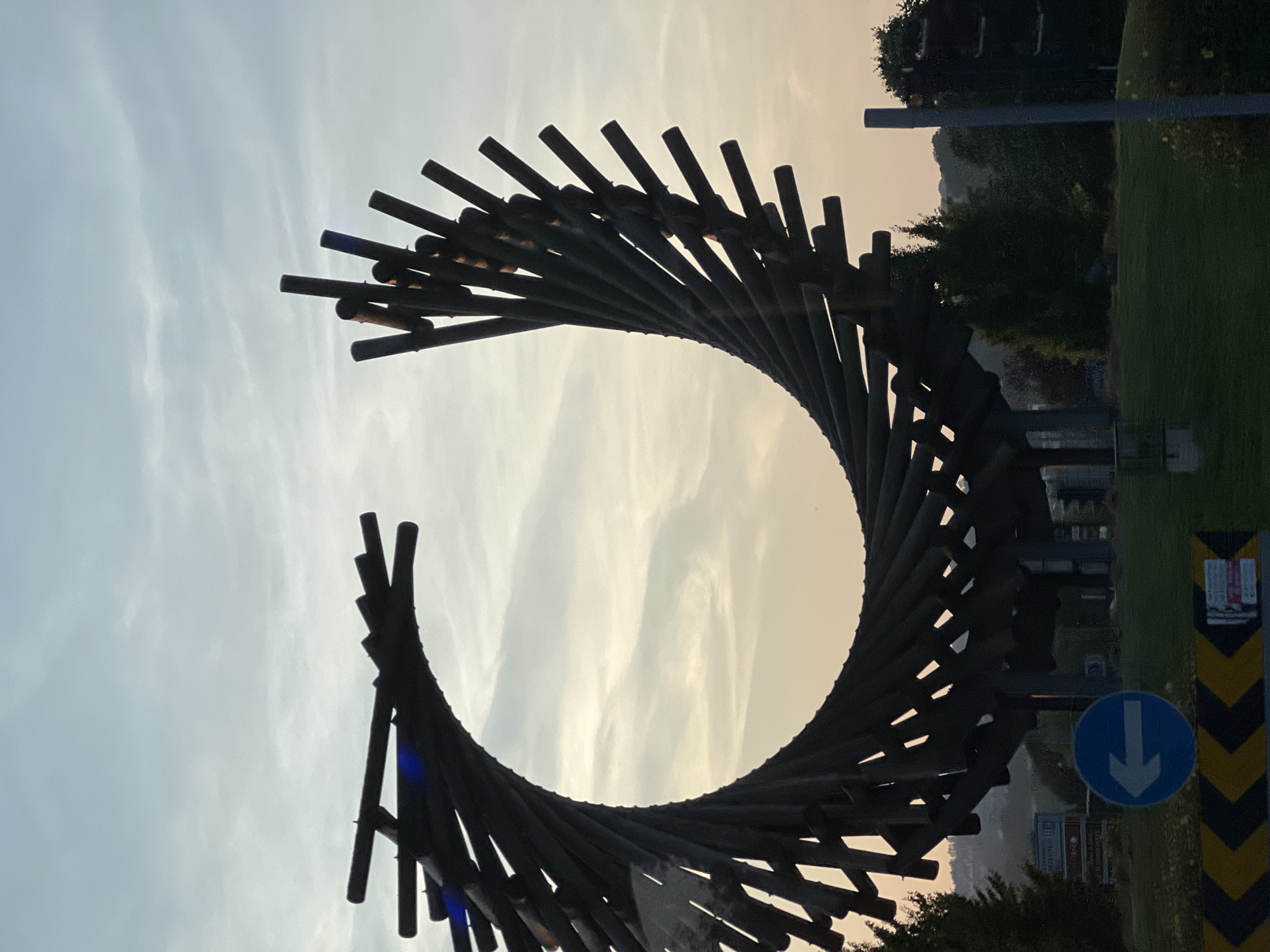 An abstract metal sculpture resembling a partially open ring or spiral is situated in a grassy area. The sculpture is composed of numerous dark, rod-like elements arranged in a twisted, helical pattern. The sky behind the sculpture is overcast with wispy clouds and displays subtle tones of grey and pale orange, suggesting either dawn or dusk. In the foreground, there is a road with a directional sign indicating a left turn. Additional elements include trees, distant buildings, and a traffic light pole. The sculpture is located at the Letterkenny Institute of Technology roundabout in Letterkenny, Ireland.