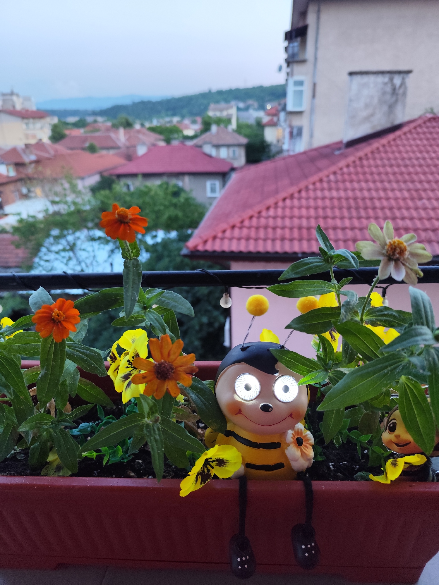 In Montana, Bulgaria, on a balcony overlooking a cityscape with terracotta roofs and residential buildings, a burgundy planter box is filled with vibrant orange and yellow flowers. Two decorative bee figures are positioned among the foliage. The larger bee, with wide, illuminated eyes, sits to the left, holding a small flower. A smaller bee figure peeks out from behind the greenery on the right. The overall scene captures a peaceful, charming setting with a touch of whimsy.