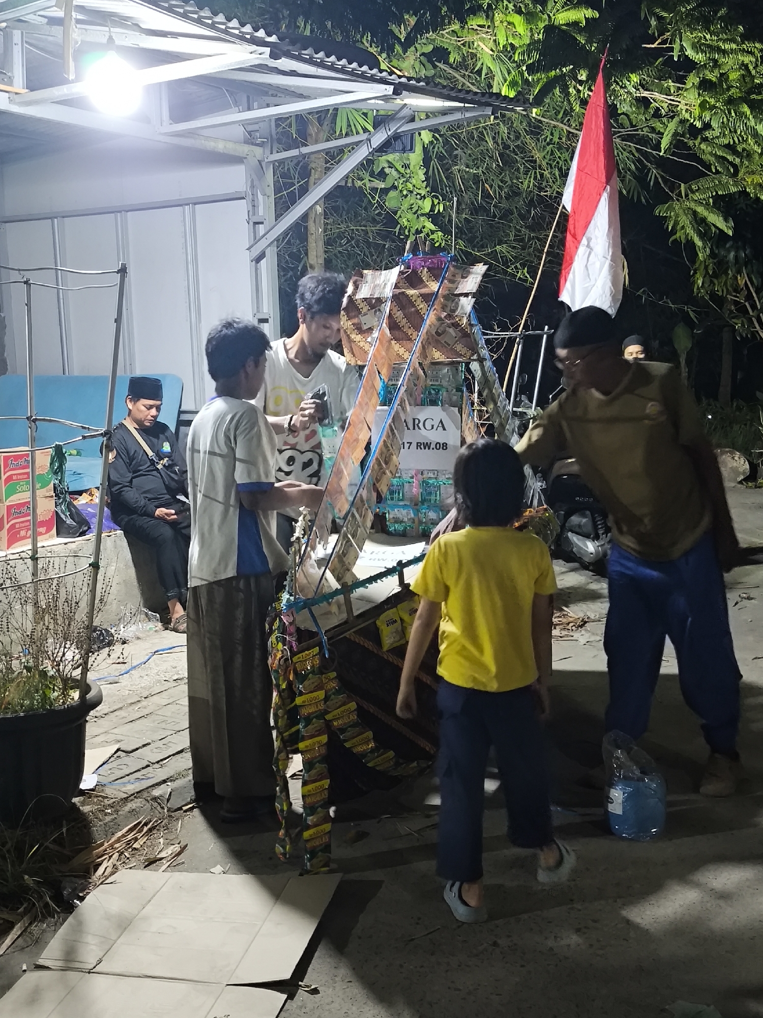 The image depicts a street-side scene at night in Cilegon, Indonesia, likely near a residential area or local market. A makeshift stall is set up, adorned with an Indonesian flag, displaying various items, possibly snacks or small goods for sale, including what appear to be packets of instant noodles. Several individuals are present: a man in a black skullcap and dark clothing sits on a low concrete ledge, appearing to observe; another man in a white t-shirt stands near the stall, holding something in his hand and interacting with a young boy in a yellow t-shirt, who is facing away from the camera; a third man in a brown shirt and blue pants is engaged with the boy. Behind the stall, a motorcycle is parked. The environment appears somewhat unkempt, with discarded cardboard and debris on the ground, illuminated by overhead lighting. The overall atmosphere suggests a local commerce setting at dusk or night.