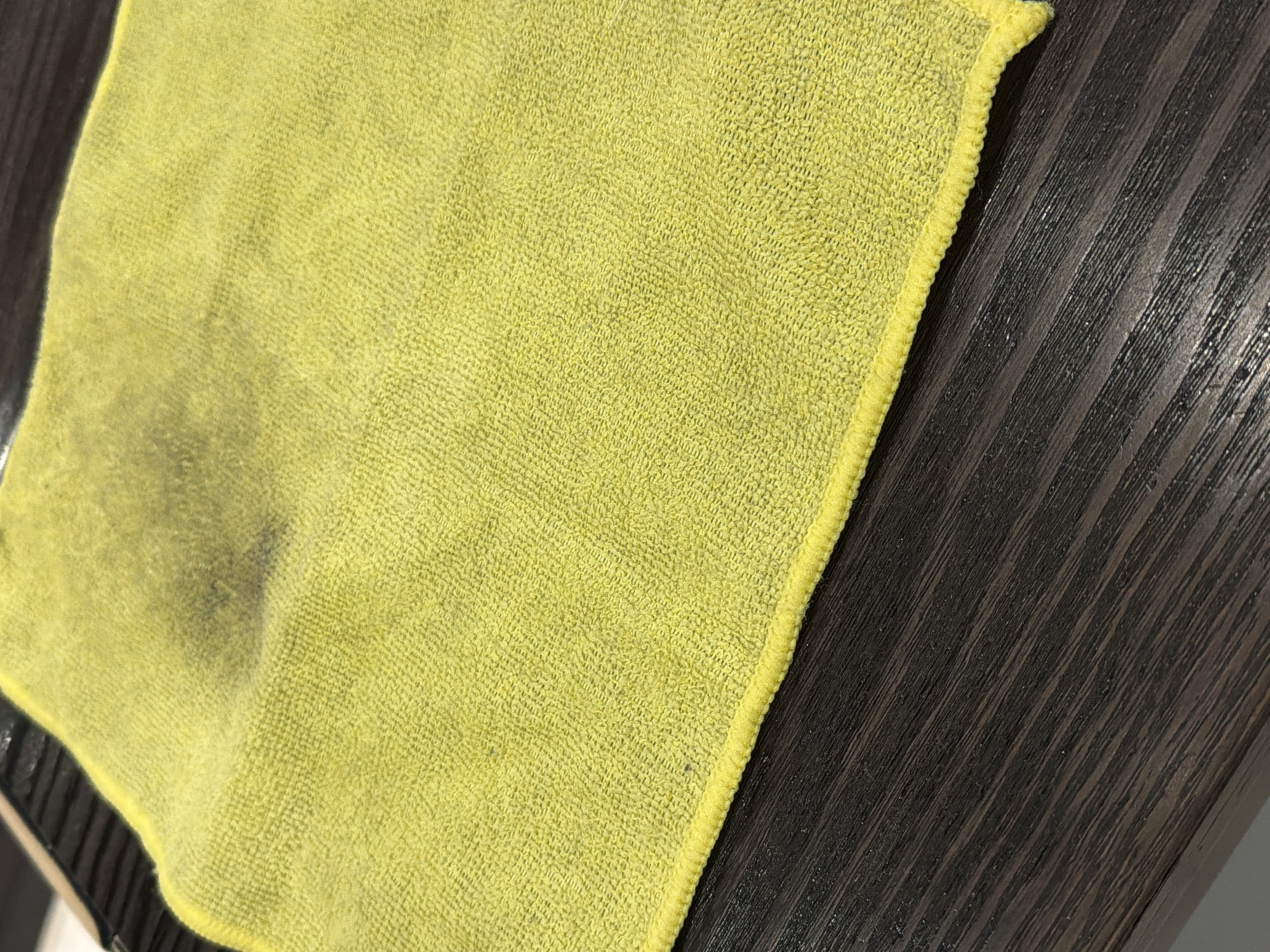 The image shows a close-up of a yellow microfiber cloth with a dark stain on the upper left portion. The cloth is placed on a dark, wood-grained surface. The lighting is overhead, casting subtle shadows. The scene is indoors, with no discernible elements indicating a specific city or location, and therefore no connection to Sofia, Bulgaria can be established.