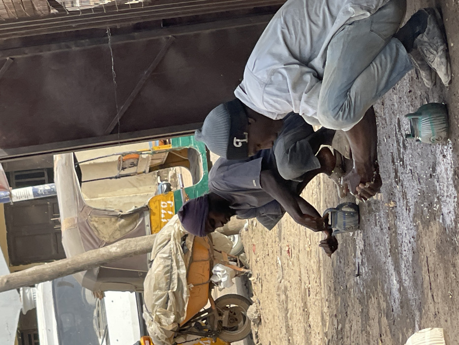 This image captures an outdoor scene in Zaria, Nigeria, featuring two adult males engaged in handwashing on an unpaved, dusty ground surface. White foamy residue and water splashes are visible on the ground directly in front of both individuals.
The man positioned on the left, with dark skin, is wearing a dark purple knit beanie, a short-sleeved grey t-shirt, and dark trousers. He is squatting, utilizing his left hand to pour water from a blue, patterned metal kettle onto his right hand, which is held cupped over the ground.
The man on the right, also with dark skin, wears a grey ribbed knit beanie with a black band featuring a white 'L' emblem, a light blue long-sleeved button-up shirt, and light-colored trousers. He is also squatting, holding his cupped hands over the ground, appearing to rinse them. A light green, patterned metal kettle rests on the ground in front of him.
In the background, a yellow and green three-wheeled auto-rickshaw (Keke Napep) with the number "1770" visible on its side is parked. A white vehicle is partially visible further to the left. A yellow wheelbarrow covered with a white tarp is situated behind the left man. A vertical wooden pole stands in the mid-ground. On the right side of the frame, a dark brown metal gate or door, secured with a wire latch, is visible. Buildings are faintly discernible in the far background. The scene is illuminated by bright daylight.