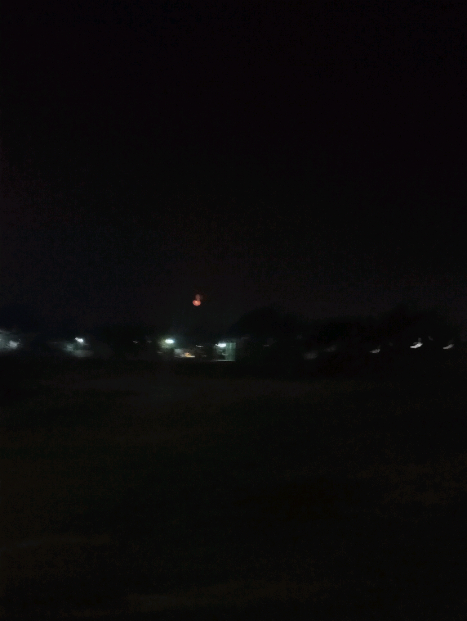 The image depicts a dark, blurry scene, likely taken at night, with a faint reddish-orange object visible in the upper central area of the sky. In the lower portion of the image, there are indistinct shapes that suggest buildings or structures with faint lights, possibly streetlights or lights from within the buildings. The ground in the foreground is dark and appears to be an open area. The overall impression is of a dimly lit, possibly rural or suburban, outdoor environment at night in Kecamatan Haurgeulis, Indonesia.
