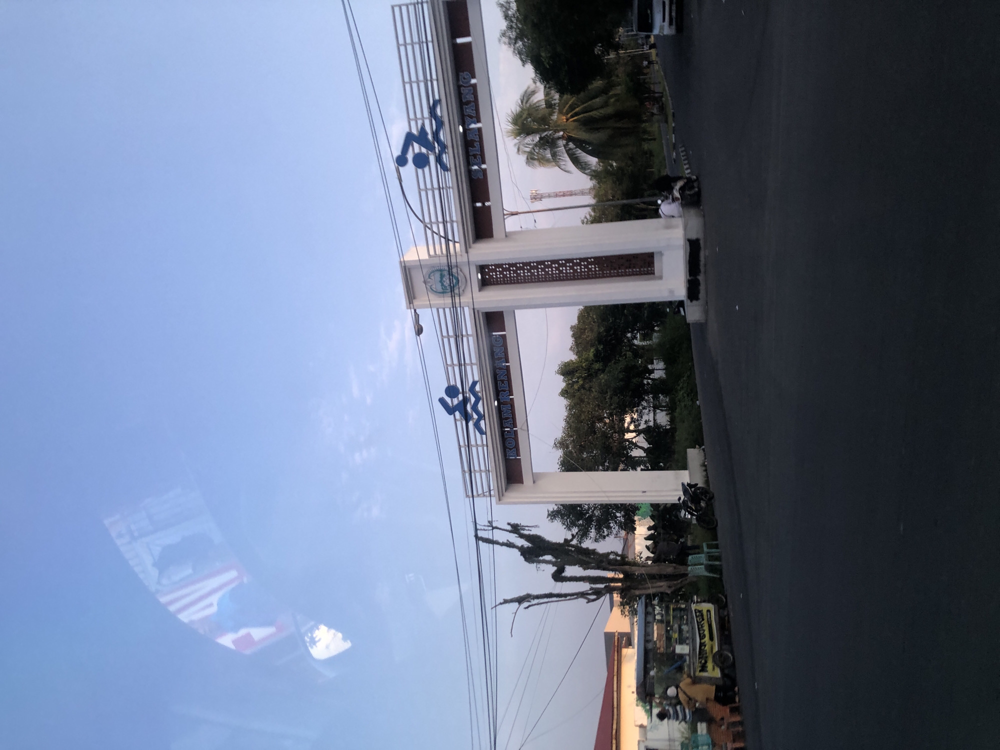 A wide-angle, eye-level shot shows an archway at what appears to be the entrance to a recreational area in the City of Medan, Indonesia, likely during the late afternoon or early morning given the lighting. The archway is white with maroon accents and features blue stylized figures of swimmers on top. Text on the arch reads "KOLAM RENANG" on the left and "SELAYANG" on the right. To the left of the archway, there's a food cart with the text "MOHON GORENG" and several people gathered around it. A dead tree with moss hanging from its branches stands prominently in the foreground, obscuring part of the food cart. In the background, there are palm trees and other vegetation. A white van is visible on the right side of the road. Power lines are strung across the sky. The sky is a clear blue with some faint clouds. There are a few motorcycles and people moving around near the archway. The scene suggests a public space in Medan, possibly near a swimming pool facility.