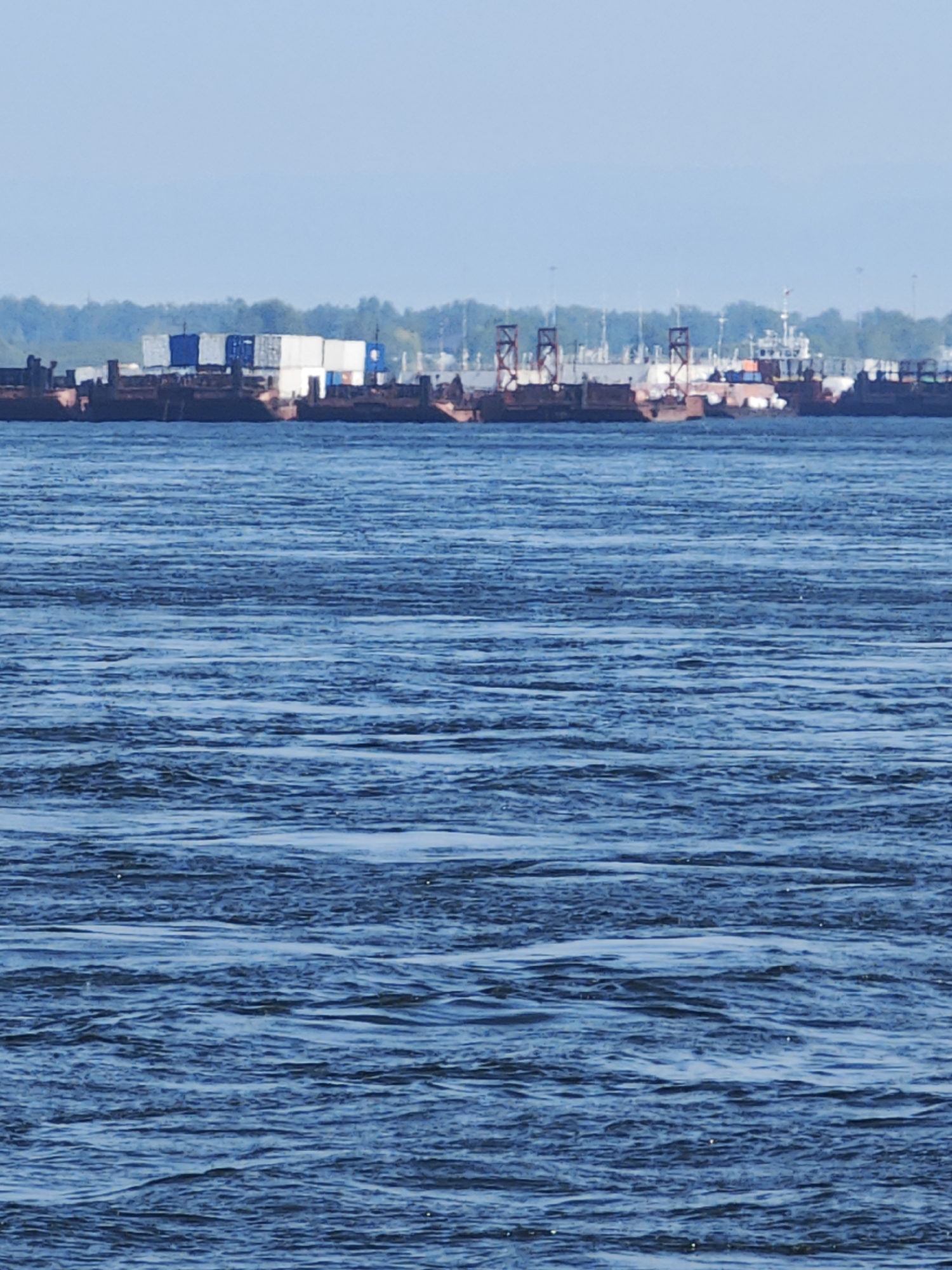 The image depicts a wide expanse of blue water, likely a river or lake, with rippling surface textures indicating movement. In the distance, along the horizon, a line of industrial barges is visible, some of which are stacked with shipping containers. The barges appear to be moored or stationary. Behind the barges, a treeline is present, suggesting a natural landscape bordering the industrial area. The sky is a pale, clear blue, with a hint of haze in the far distance. The scene appears to be an open-air environment, possibly a port or industrial waterway, on a clear day. The location is not specified to be in Krasnoyarsk, Russia, as this information is not visually discernible from the image.