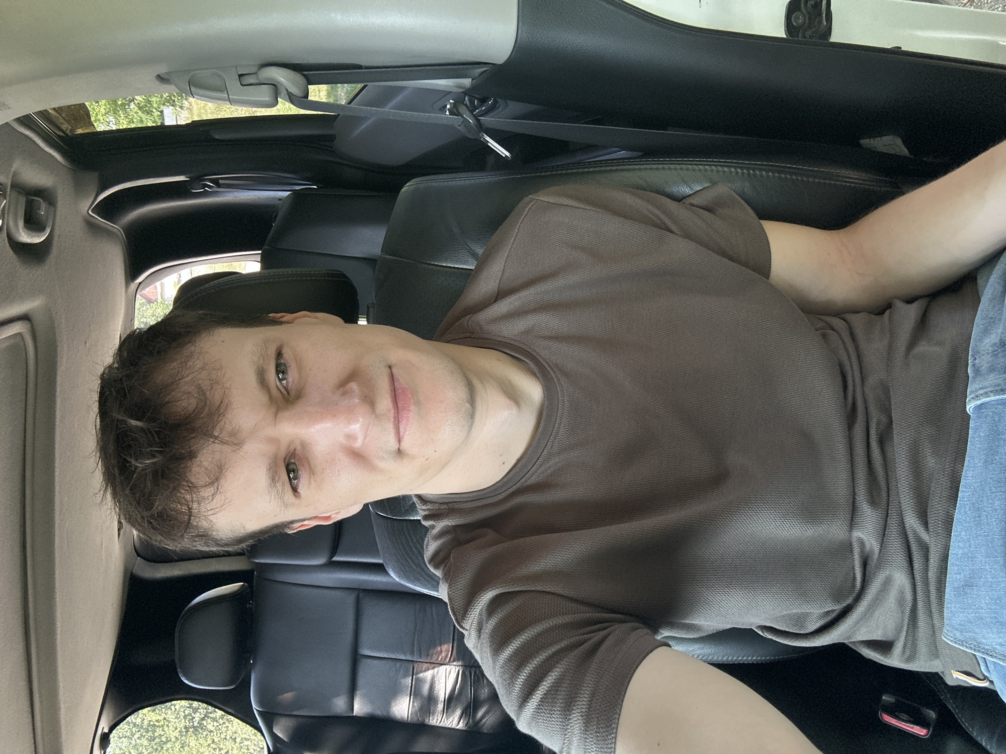The image shows a young man in the passenger seat of a vehicle, looking at the camera. He is wearing a brown t-shirt and blue jeans. The interior of the car is visible, including black leather seats and the dashboard. Sunlight filters in from outside, creating shadows on the seats. The background appears to be outdoors, with trees and greenery visible through the windows, suggesting a rural or suburban setting outside the city of Makiv, Ukraine. The man's expression is neutral.