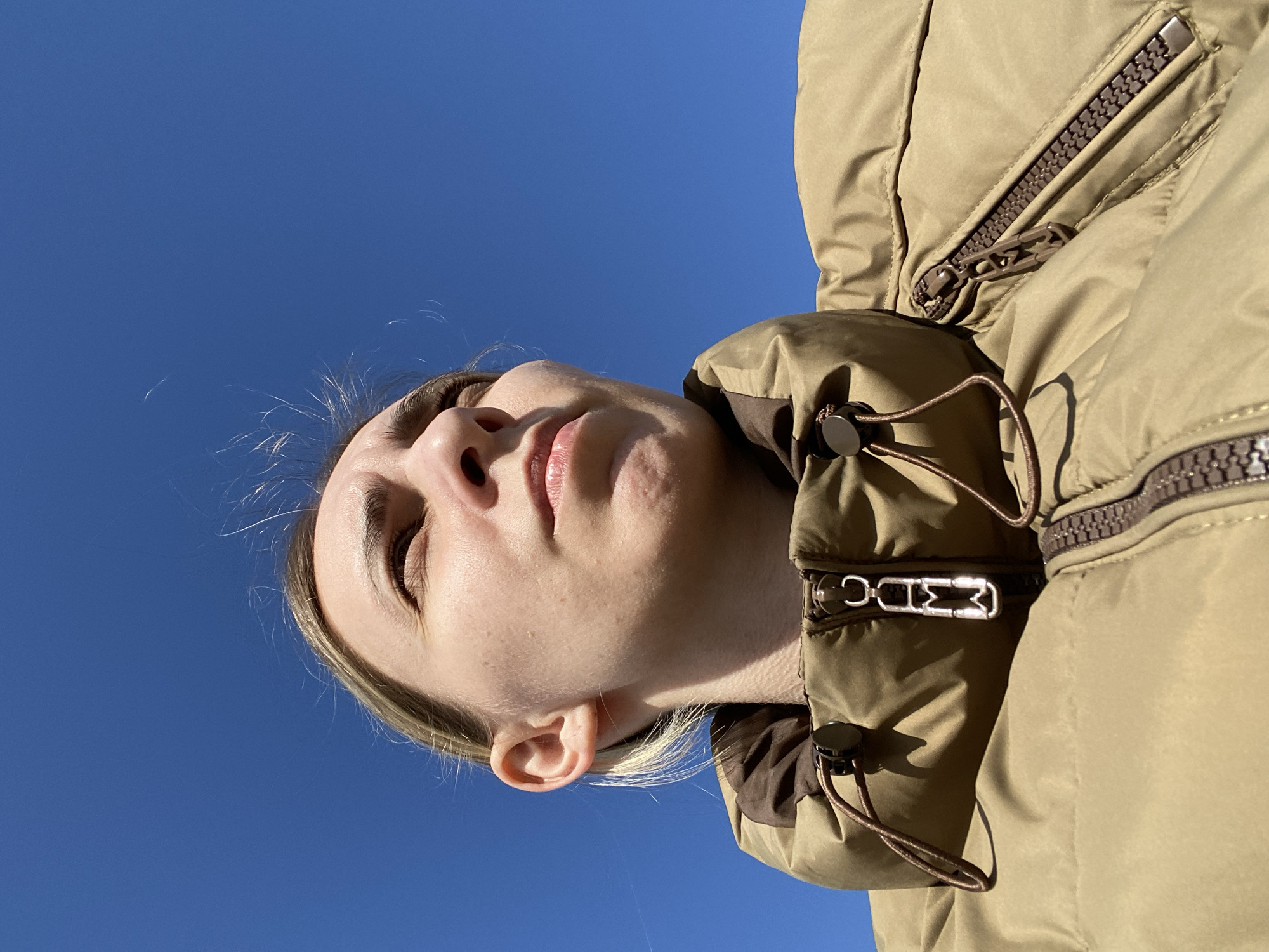 A low-angle close-up shot depicts an adult female positioned in the lower left portion of the frame, looking upwards and to the viewer's right against a clear, bright blue sky. Her blonde hair has wisps visible against the sky. Her eyes are slightly squinted, suggesting direct sunlight, which casts shadows beneath her jawline and on the folds of her clothing. She is wearing a tan or khaki-colored quilted puffer jacket with a brown inner collar. The jacket's main metallic zipper features a distinctive geometric pull-tab. A brown drawstring with a plastic toggle is visible on the jacket's collar, and a brown plastic zipper with a textured pull-tab is visible on a pocket to the right. The background consists solely of an unblemished blue sky. The specific location in Chaikovsky, Russia, cannot be visually confirmed from the image content.