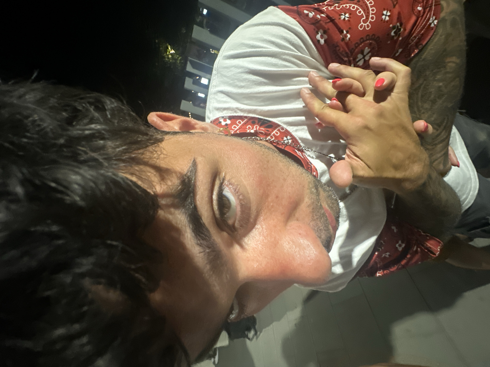 A close-up, high-angle, dimly lit outdoor shot shows a young person with dark, messy hair, brown eyes, and light skin. They are wearing a white t-shirt with a red bandana-like pattern on the sleeves and collar. The person's hands are clasped together, and their fingernails are painted bright pink. A silver chain with a pendant is visible around their neck. The background features dark shadows, a hint of a light-colored railing, and some blurred lights in the distance, suggesting it is nighttime. The ground appears to be paved. The image does not provide enough information to determine the specific location in El Masnou, Spain.