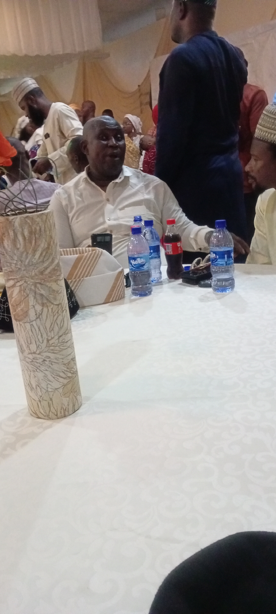 In Gandu Albasa, Nigeria, a group of people are gathered at a white-clothed table, where several bottles of water and a bottle of Coca-Cola are placed. In the foreground, a man wearing a white shirt is seated, looking directly at the camera with a surprised expression. Behind him, a man in a navy blue shirt stands, while other individuals, some in traditional attire and headwear, are visible in the background, suggesting a social gathering or event. The environment appears to be an indoor venue decorated with light-colored drapery.