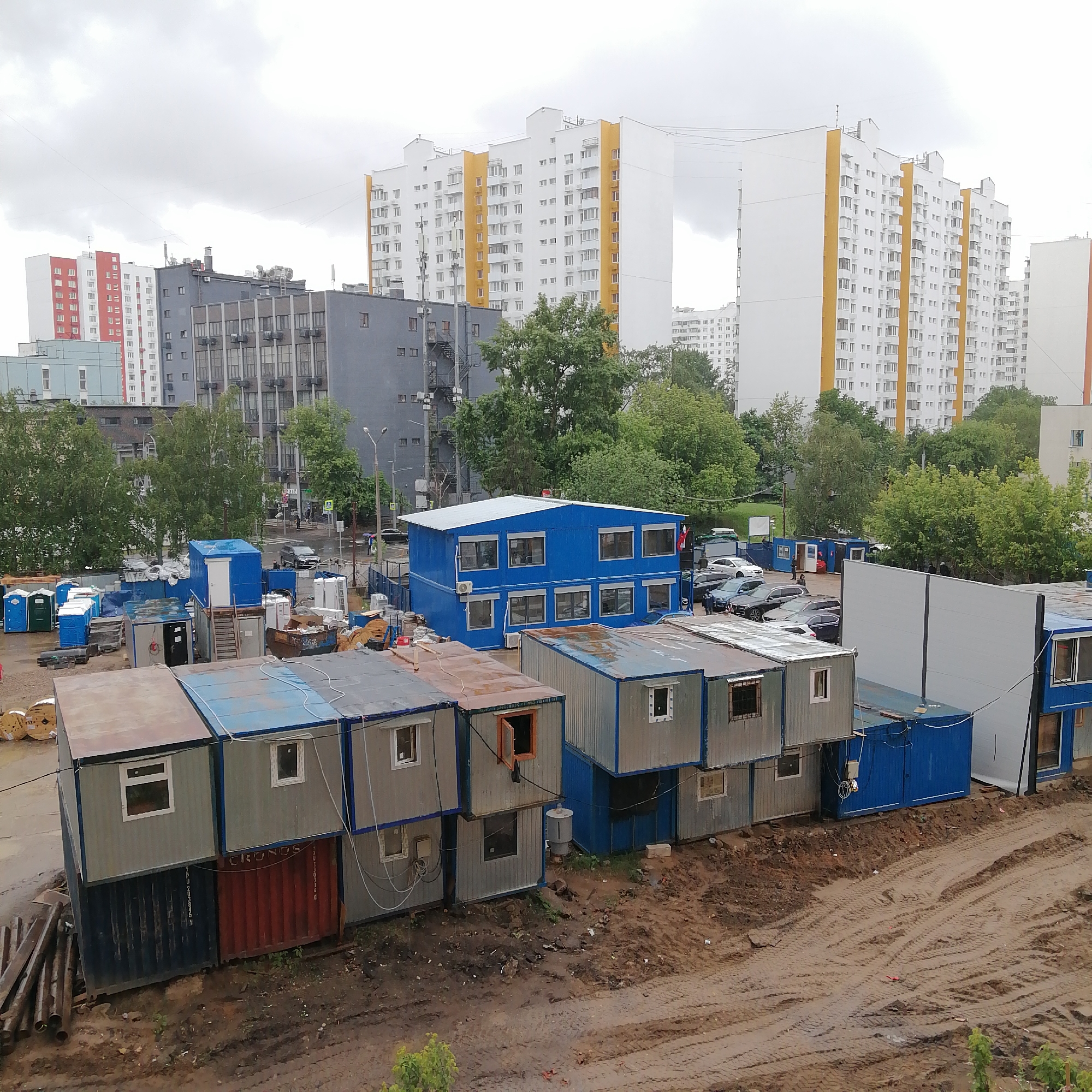 The image depicts a construction site or temporary office area located in an urban setting. Dominating the foreground are several stacks of portable modular buildings or shipping containers, which appear to serve as temporary housing or office spaces. The containers exhibit a range of colors, including silver, blue, and red, and show signs of wear and tear.
In the background, there are high-rise residential buildings, some of which are white with yellow accents and others in shades of gray and red. These suggest that the construction site is situated in a densely populated area of Unknown, Unknown.
There are visible tire tracks in the muddy ground, suggesting recent vehicle activity. Other portable facilities, such as portable toilets, are scattered around the site. Several cars are visible. Overhead, the sky is overcast, indicating cloudy or rainy weather conditions.